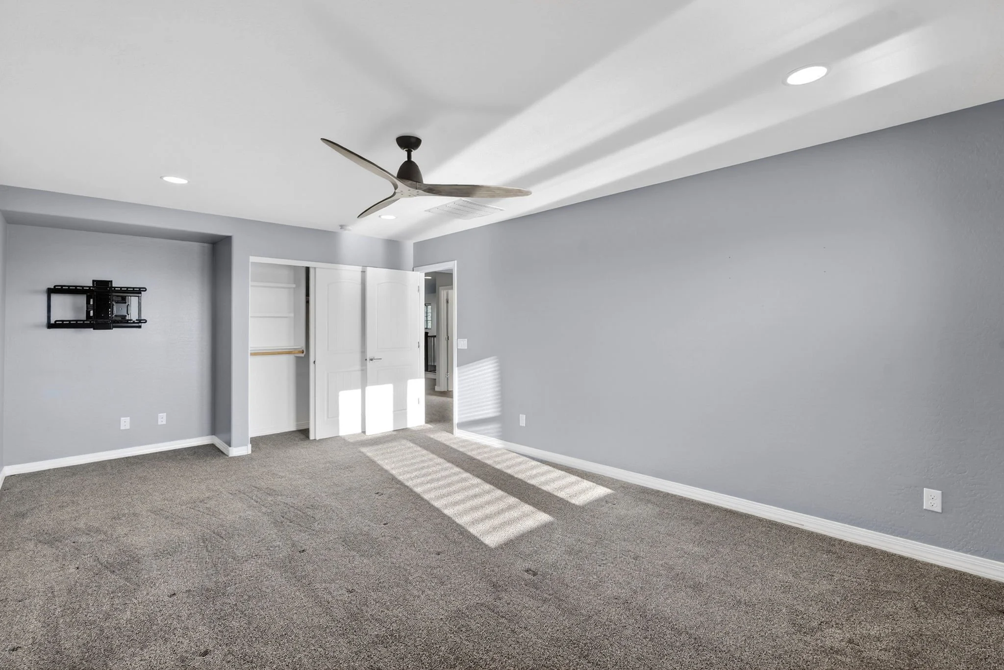 Empty room with gray walls, carpeted floor, ceiling fan, and a wall-mounted TV bracket.