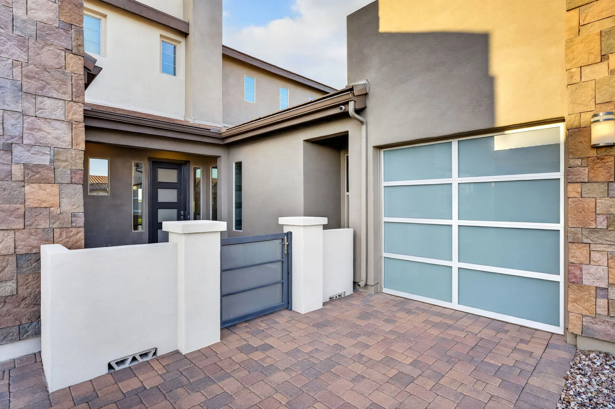 The image shows the exterior of a modern house with a combination of stone and stucco walls. There is a garage with a blue and white segmented door, a black front door with vertical window panes, and a small front yard area with brick paving and a lo