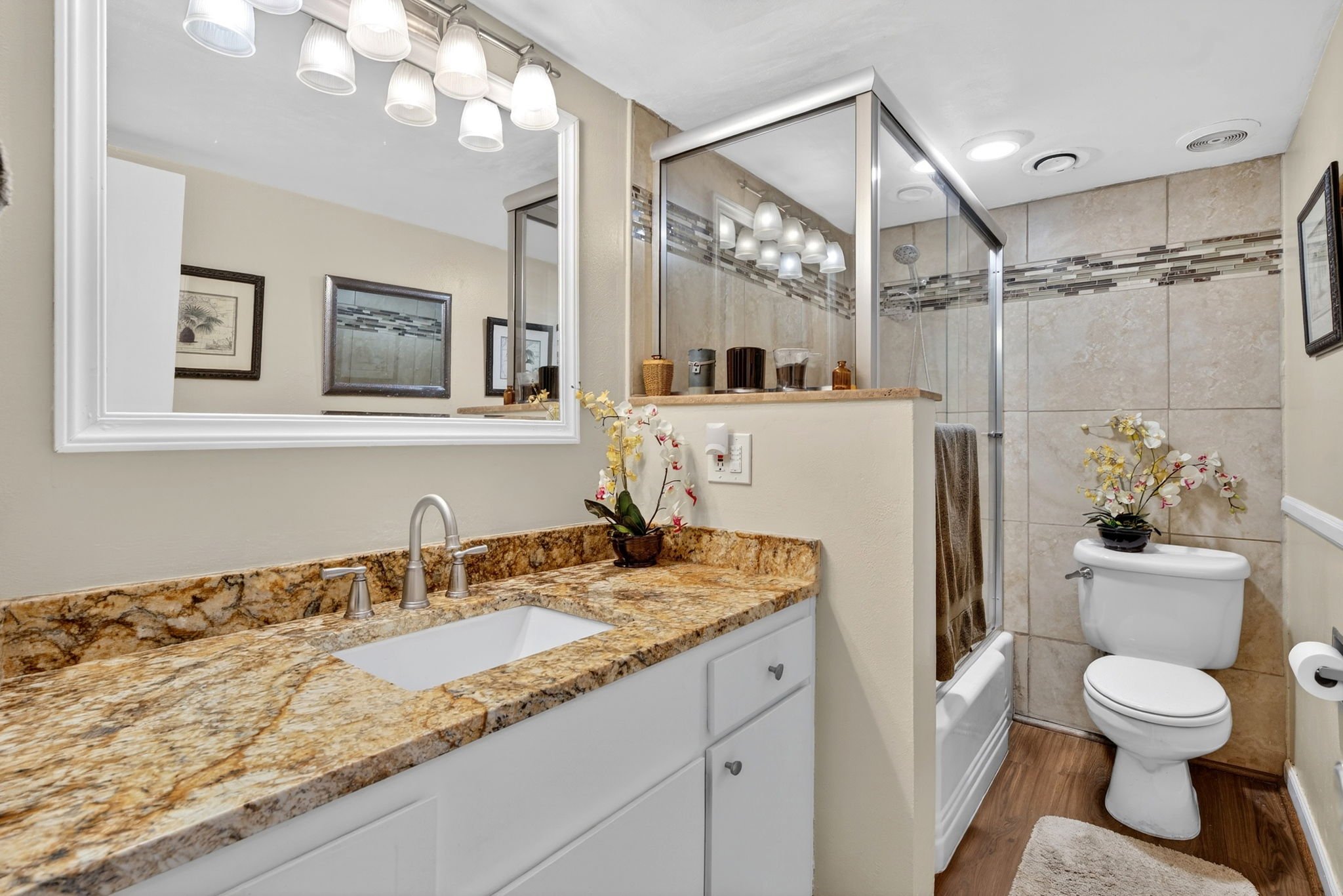 A bathroom with a granite countertop vanity, mirror, and a glass shower enclosed with tile walls. There are framed pictures on the wall and a decorative flower arrangement on the counter.
