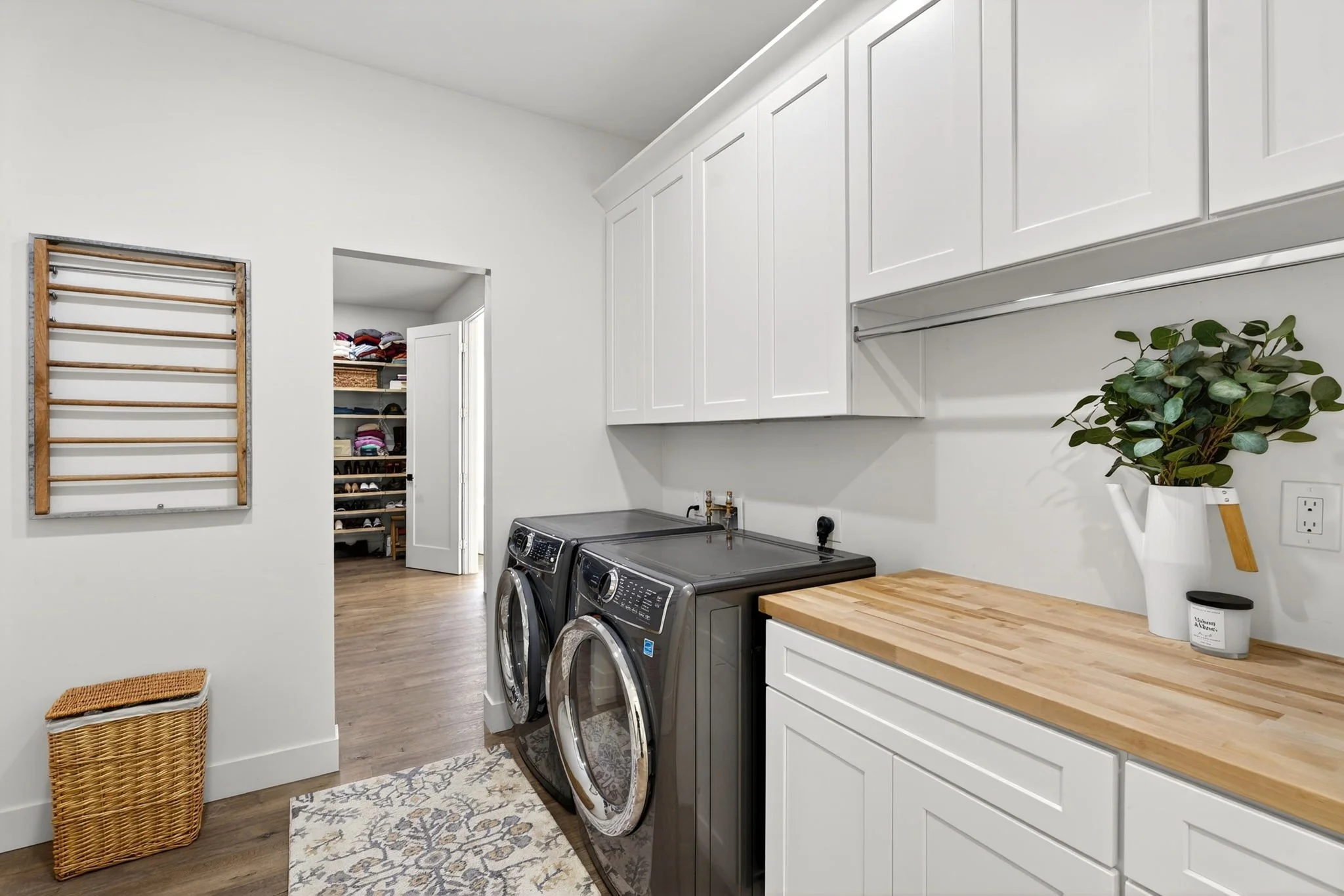 Laundry room with two front-loading washers, white cabinets, a wooden countertop, a potted plant, and a laundry basket.