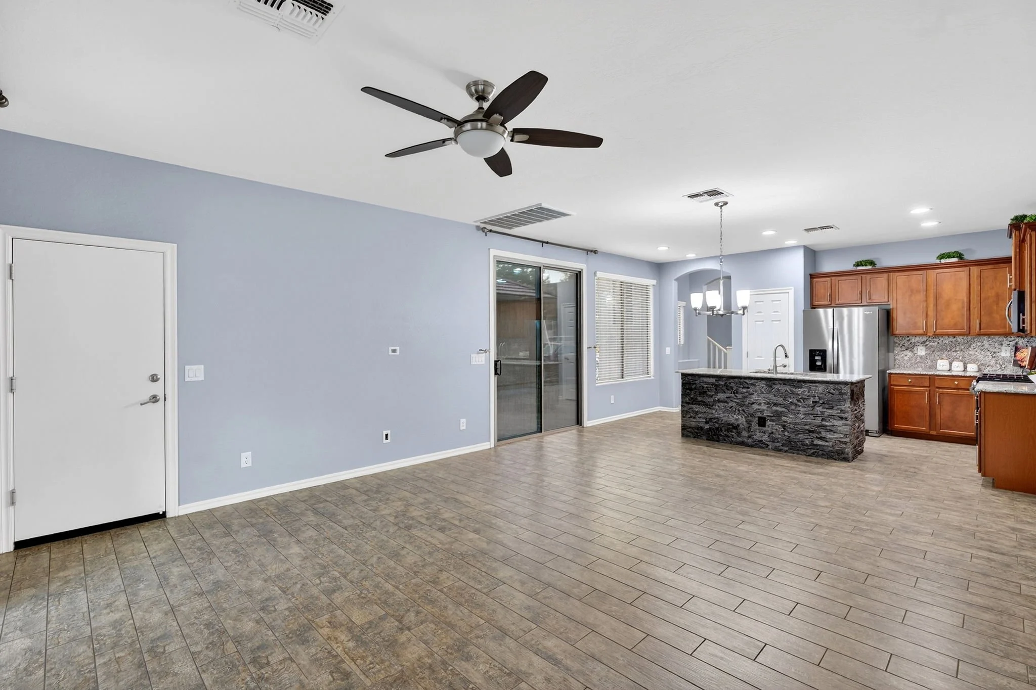 Interior view of a modern kitchen and living area with light blue walls, wooden flooring, a ceiling fan, and a kitchen with wooden cabinetry, granite countertops, and stainless steel appliances.