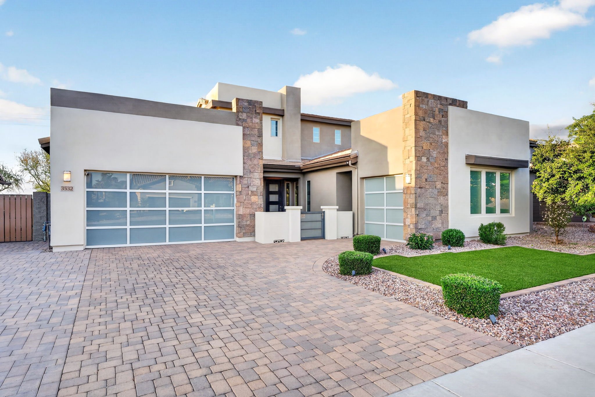 Front view of a modern house with a stone and stucco exterior, large garage door, small front yard with bushes, and a brick driveway.