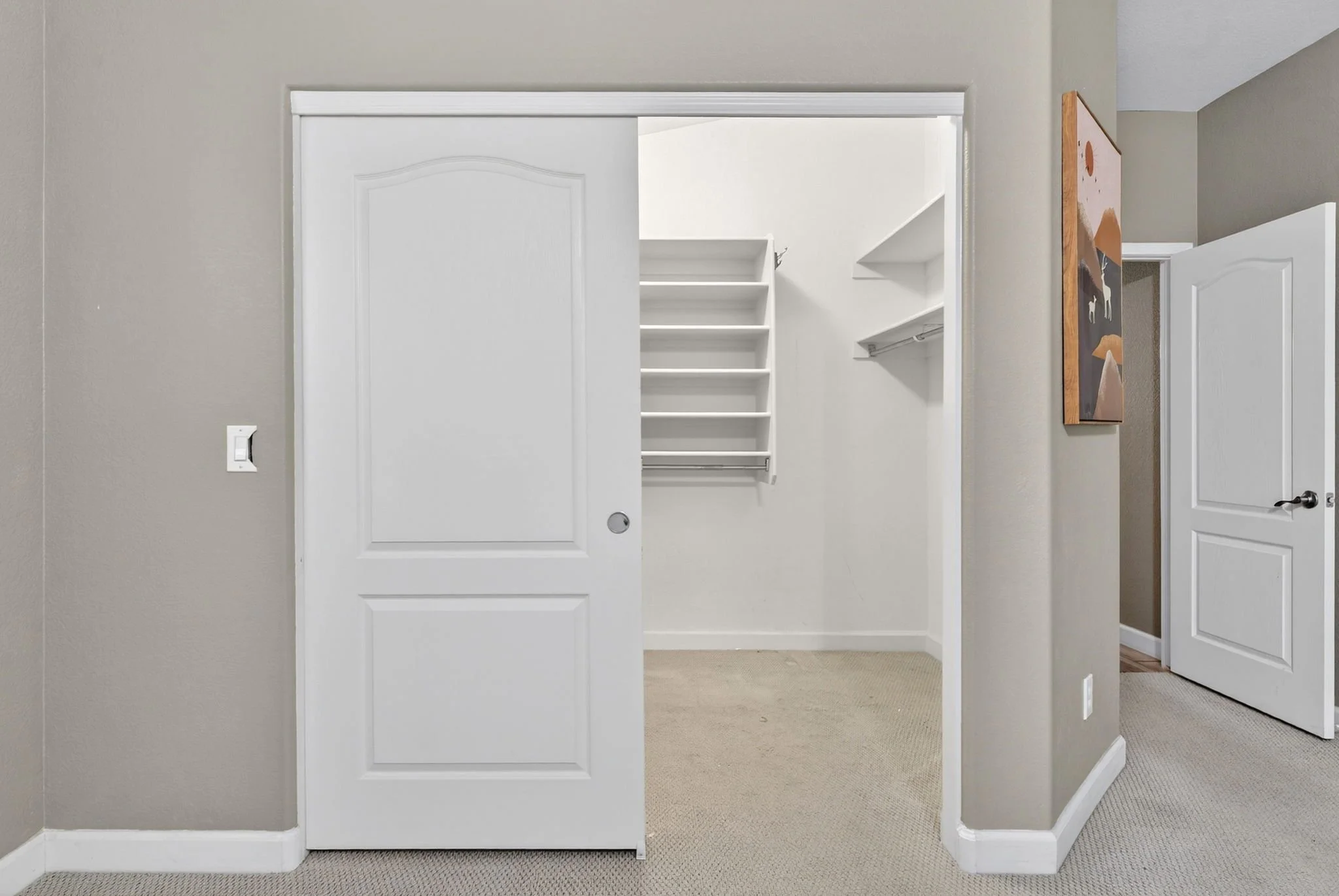 A room with white sliding closet doors, one of which is partially open revealing white shelves inside, and beige carpet flooring. A painting or picture hangs on the wall outside the closet.