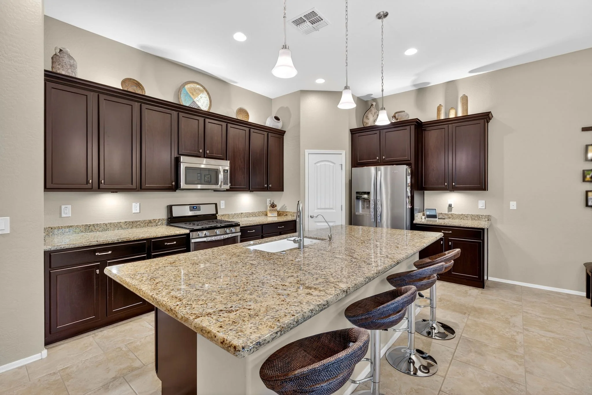 Kitchen with granite countertops, dark wood cabinets, stainless steel appliances including a refrigerator and microwave, beige tile floor, and bar stools lined up at the counter. Decorative vases and plates on top of the cabinets, pendant lights hang