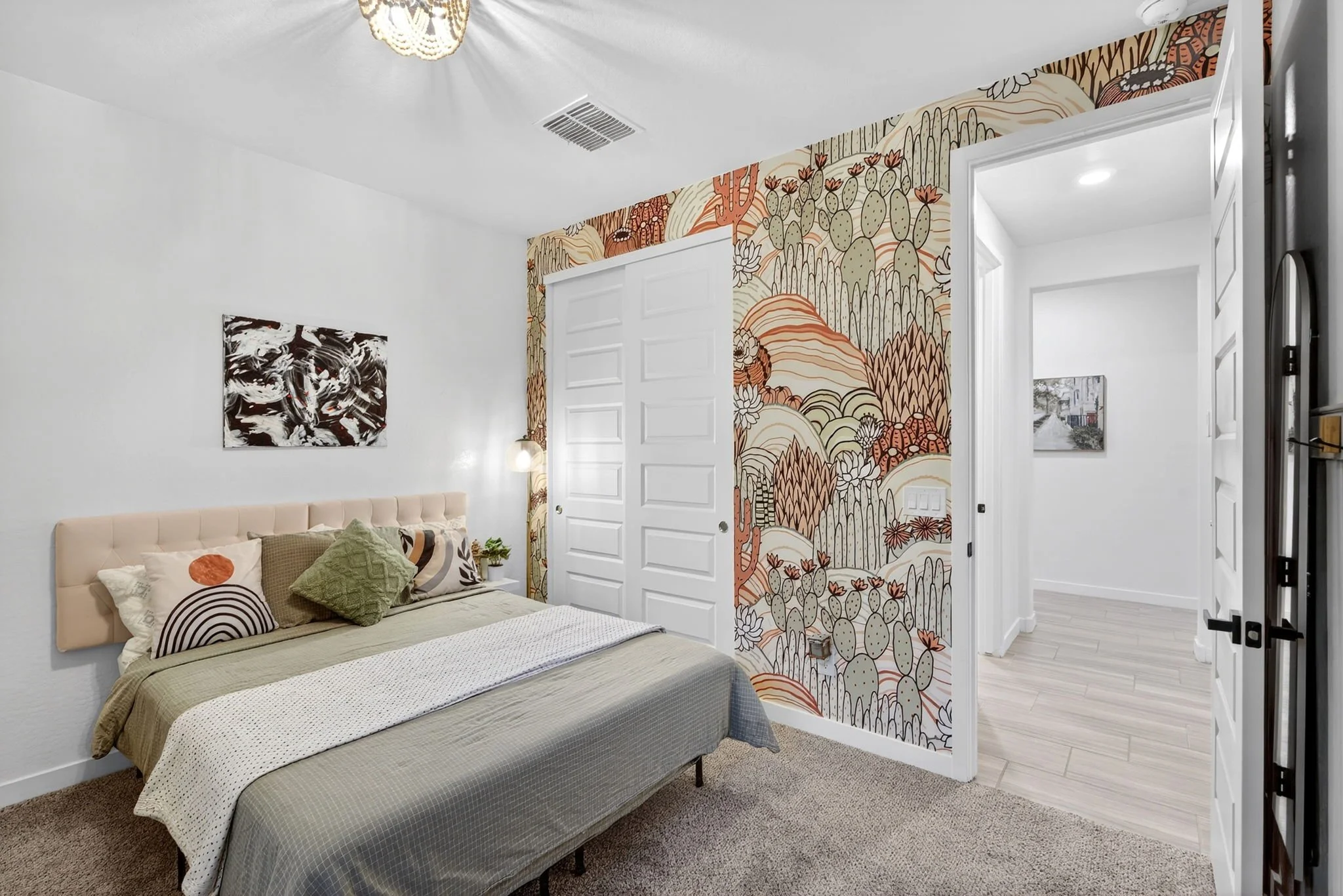 A bedroom with a bed, decorative pillows, and a nightstand. An artistic wall with desert theme wallpaper is behind the bed, with a white door and artwork on the opposite wall.