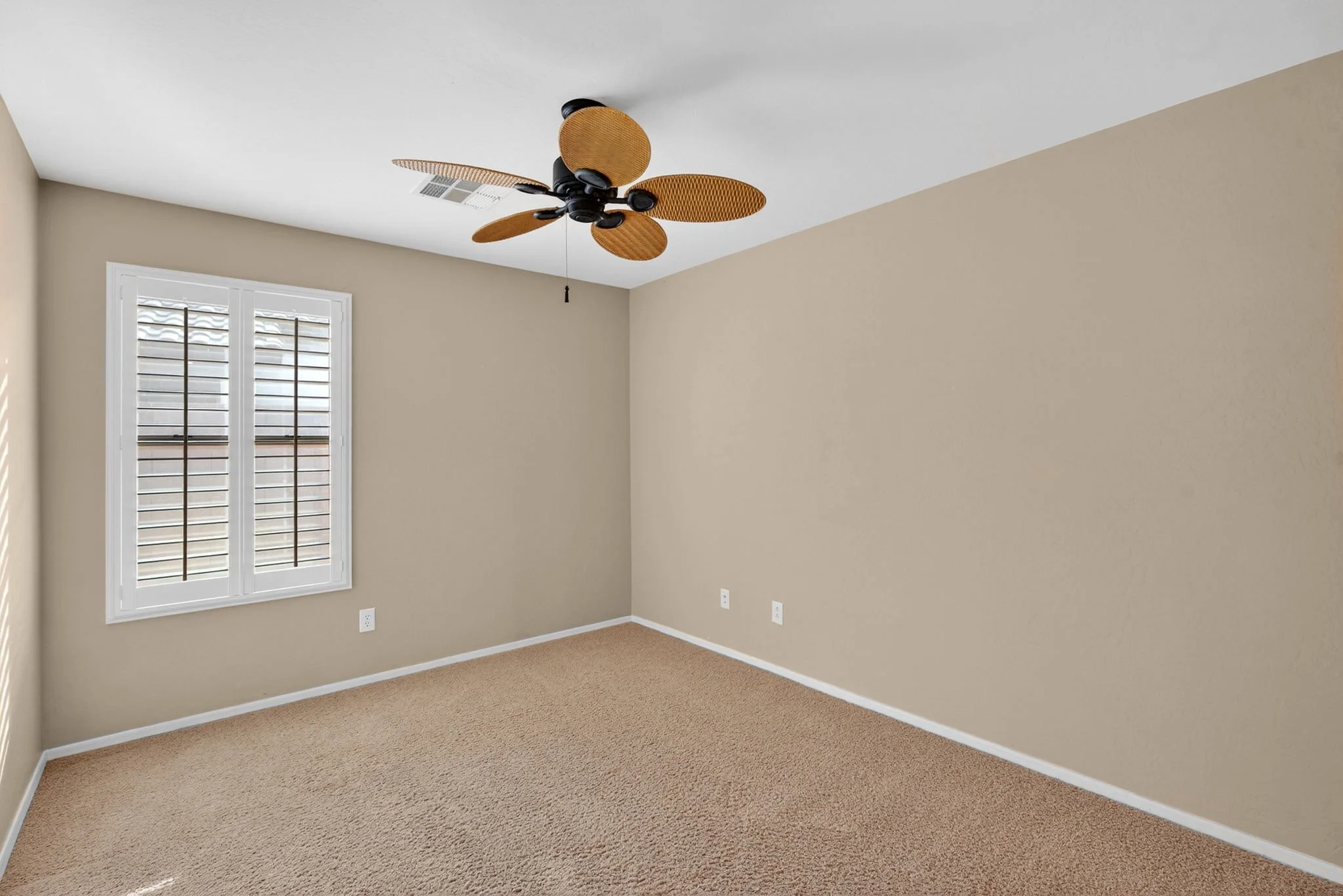 Empty beige-colored room with a window with white plantation shutters, a ceiling fan with brown blades, and beige carpeting.