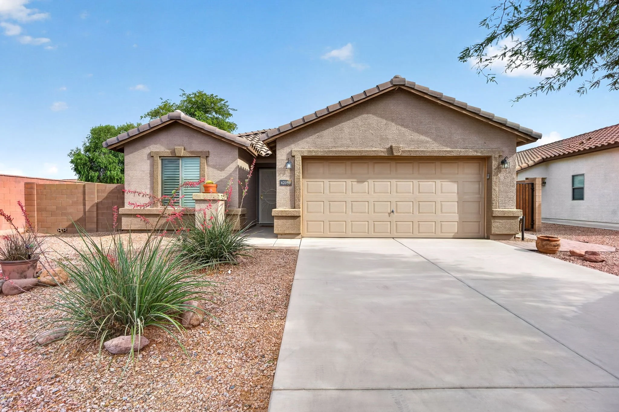 Front view of a beige-colored house with a closed garage door, a front door with a small window, desert landscaping, a concrete driveway, and a clear blue sky.