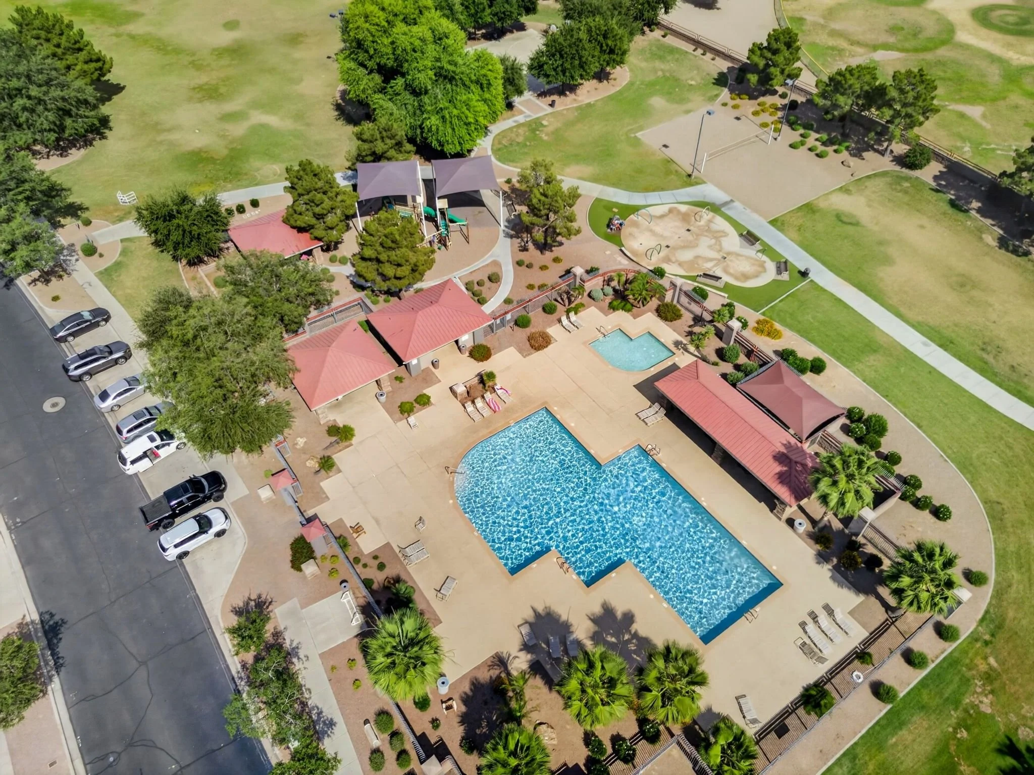 An aerial view of a community park featuring a playground with slides and swings, a swimming pool, tennis courts, walking paths, trees, and parked cars along the street.