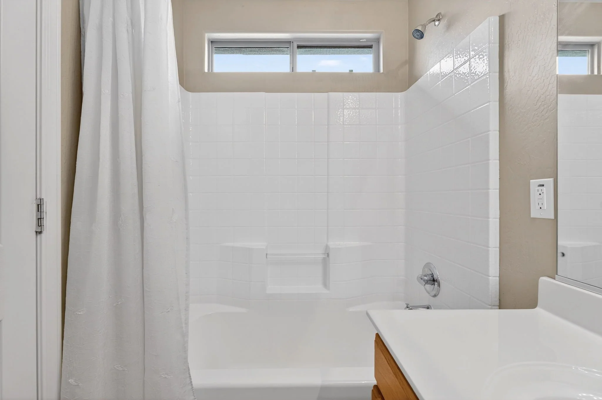 Bathroom with a white bathtub, showerhead, white tiles on the wall, a window above the bathtub, and a mirror reflecting part of the room.