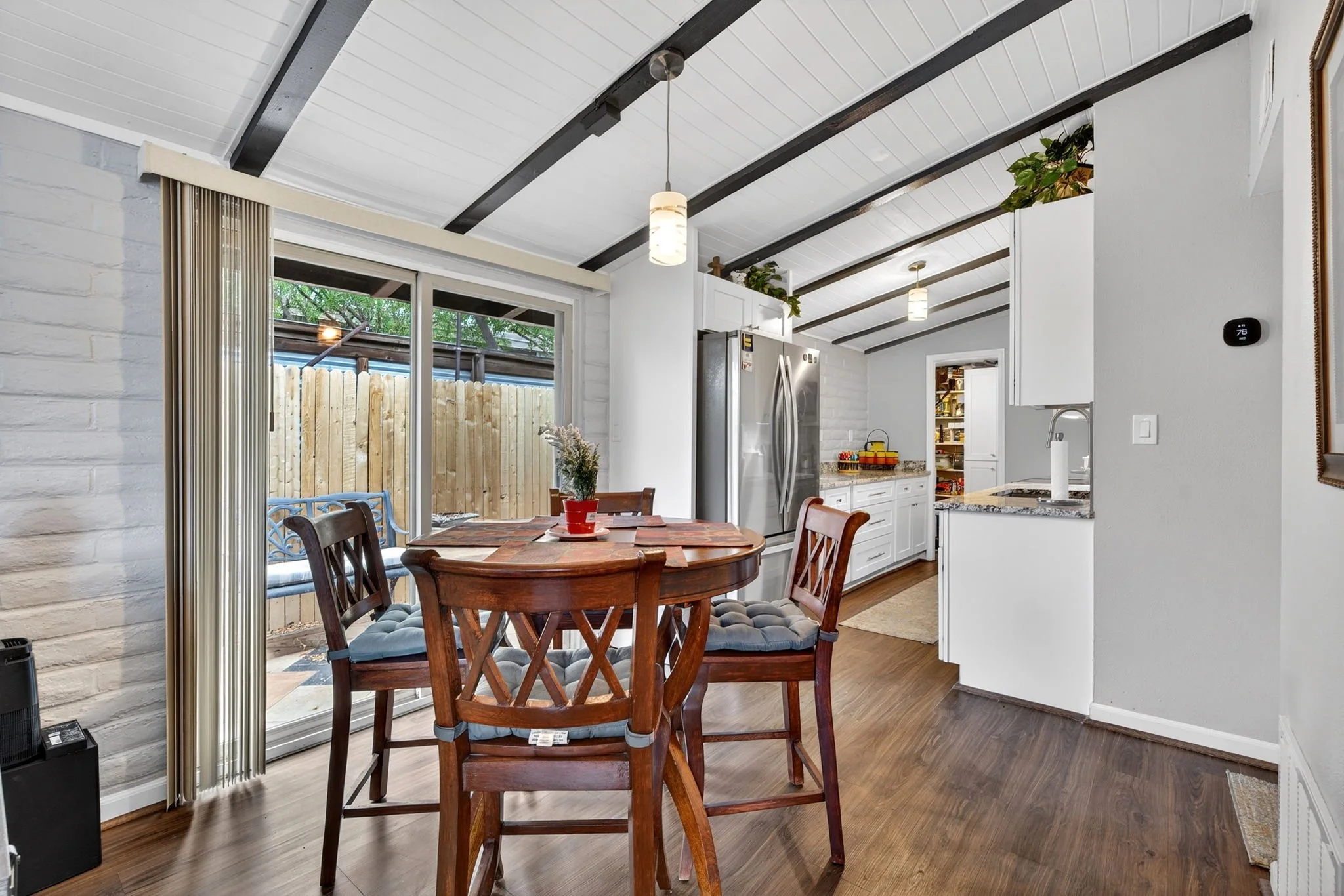 A dining area in a kitchen with a wooden table, chairs, and a window leading outside. The kitchen has white cabinets, a stainless steel refrigerator, and ceiling lights. There are some indoor plants on the ceiling beams.