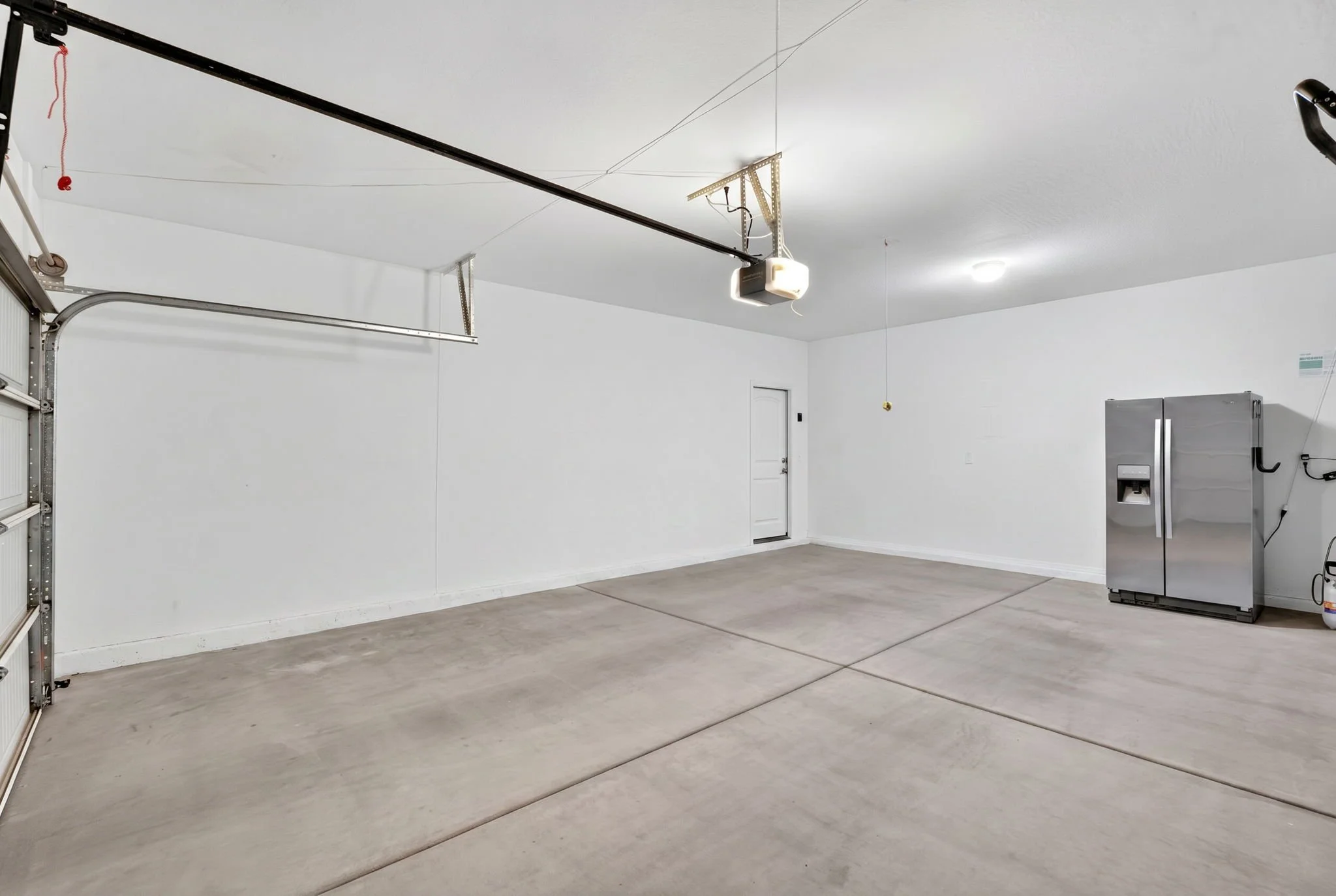 Empty garage with a concrete floor, white walls, a closed garage door, a ceiling-mounted garage door opener, a closed white door, and a stainless steel refrigerator in the corner.
