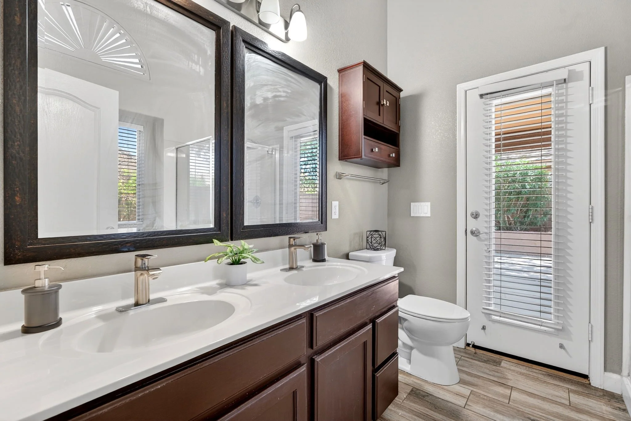 A bathroom featuring a double sink vanity with dark wood cabinets, a large mirror with a dark frame, a toilet, and a glass door leading outside with blinds. There is a small potted plant, soap dispenser, and a decorative object on the vanity. The wal