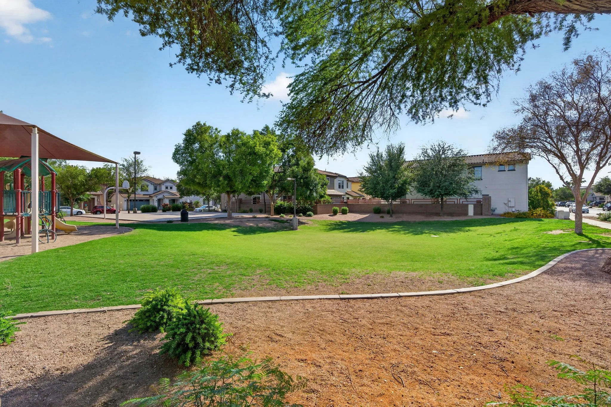 A green park with grass, trees, and a playground. Residential houses and a parking lot are visible in the background under a blue sky with some clouds.
