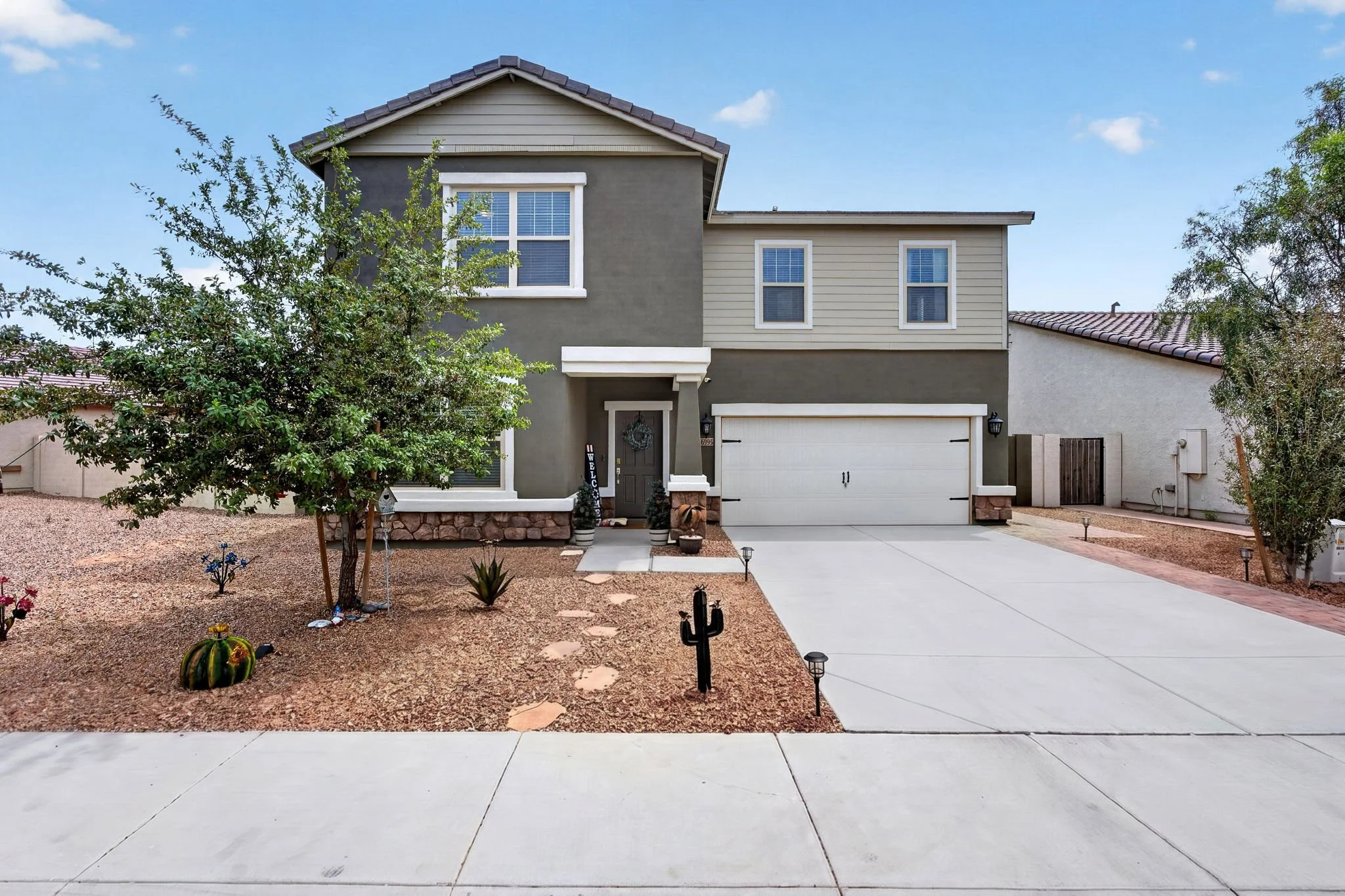 A two-story house with a gray front and a beige side, a white garage door, and a front porch with plants and decorations. The front yard is landscaped with gravel, small plants, and cactus, and a concrete driveway leads up to the garage. The sky is c