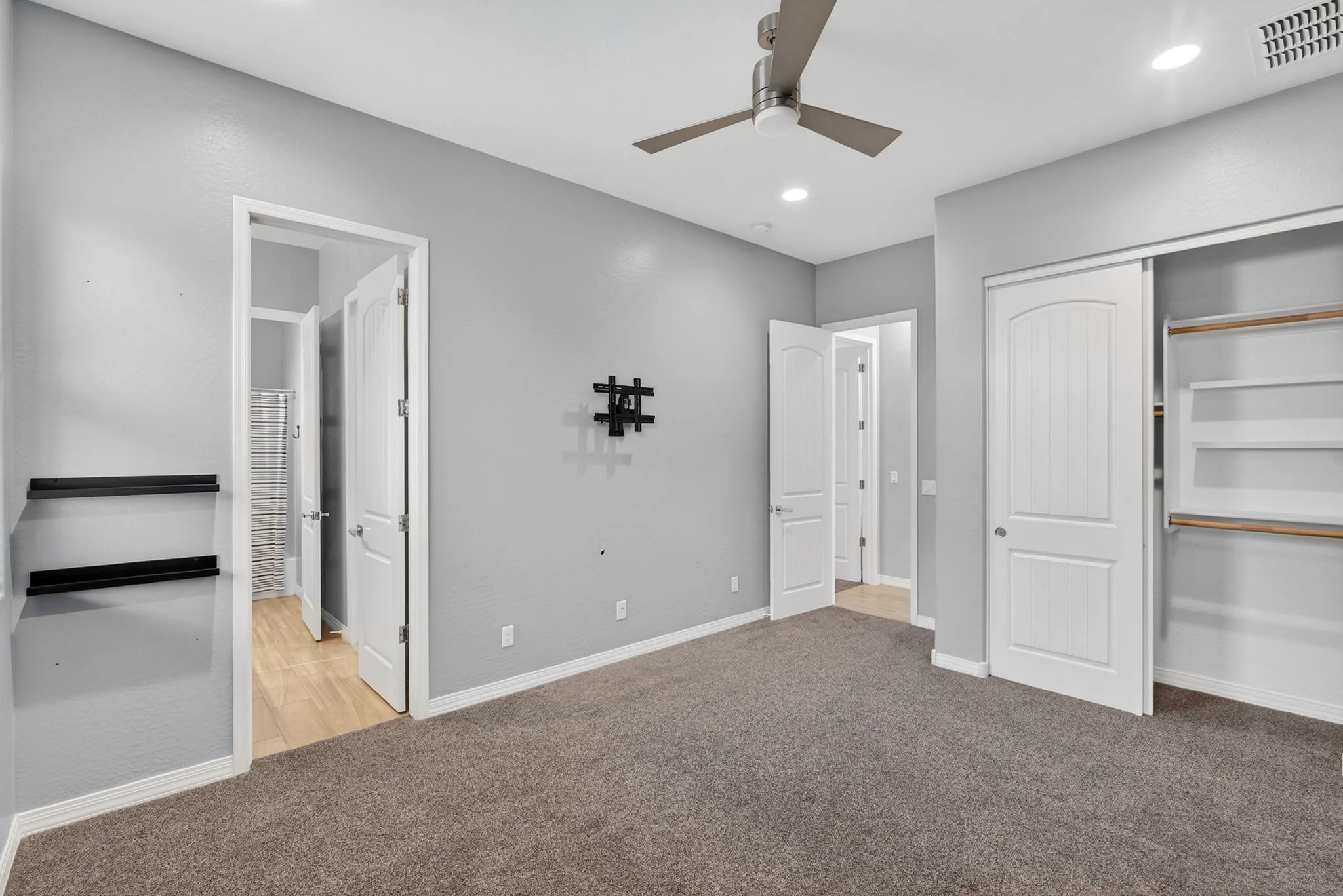 Interior view of a bedroom or living space with gray walls, white trim, a ceiling fan with lights, a closet with sliding doors, and multiple open doors leading to other rooms. The floor has a combination of hardwood and carpet.