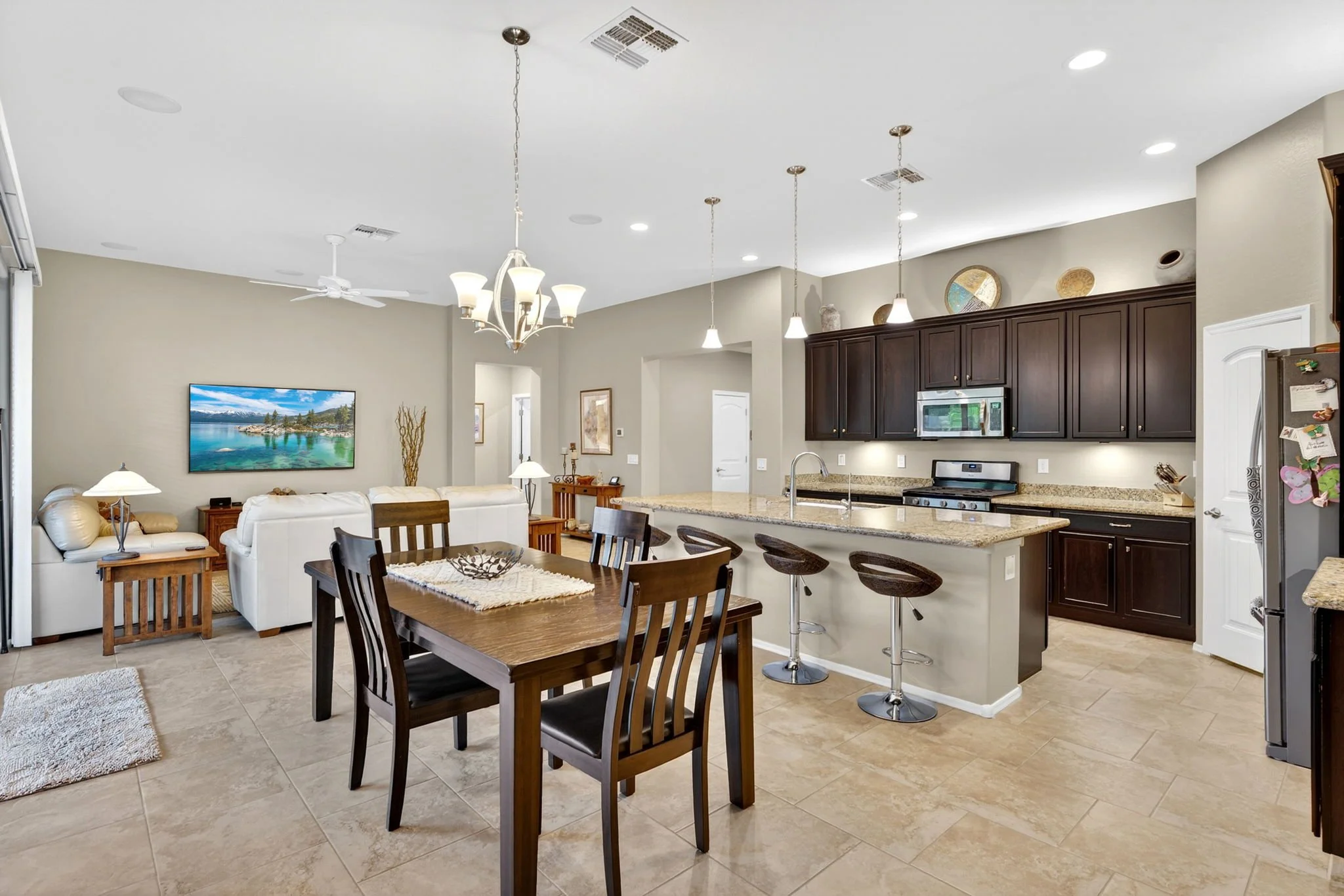 Open-concept living room and kitchen with dark wood cabinets, granite countertops, pendant and chandelier lighting, a wall-mounted TV, beige walls, ceiling fan, and tiled flooring.