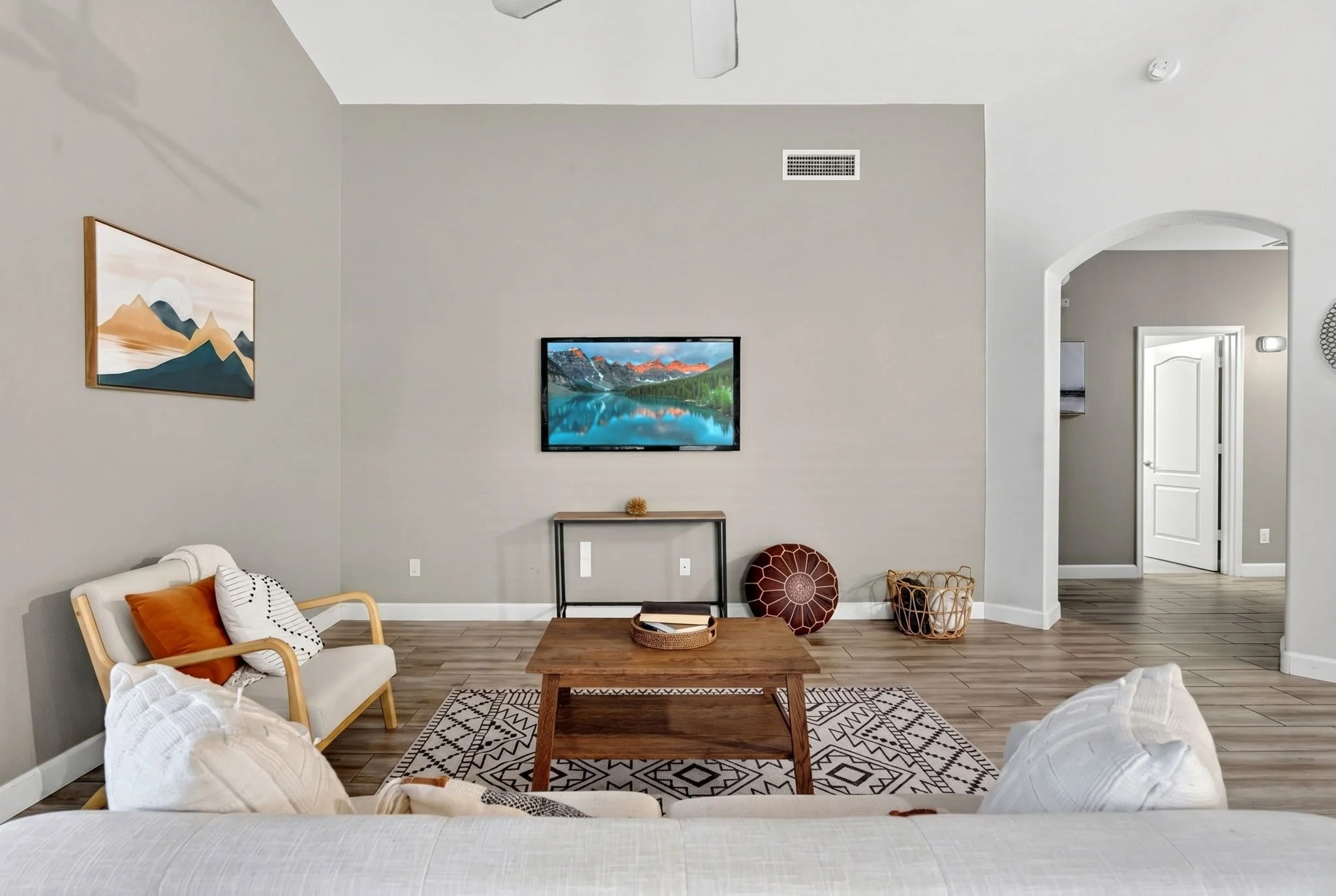 Living room with a wall-mounted TV, a wooden coffee table, a patterned rug, a white cushioned armchair, and decorative pillows, with artwork on the wall and open doorway leading to another room.
