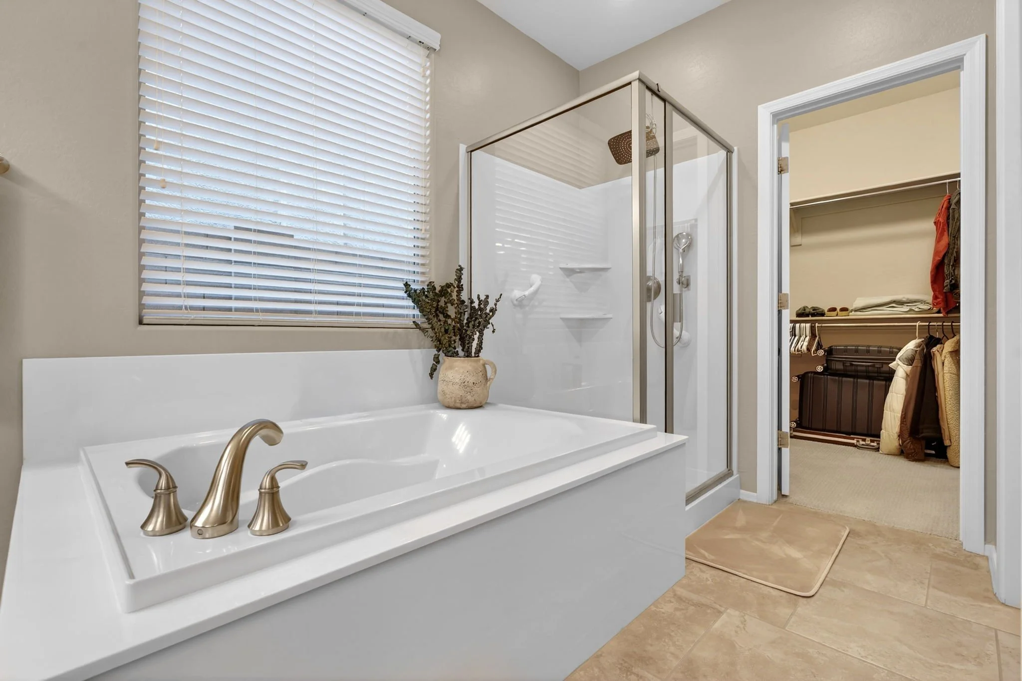 A bathroom with a white bathtub, gold fixtures, a window with white blinds, a plant in a beige pot, a shower with glass enclosure, and an open walk-in closet with clothing and a suitcase visible.