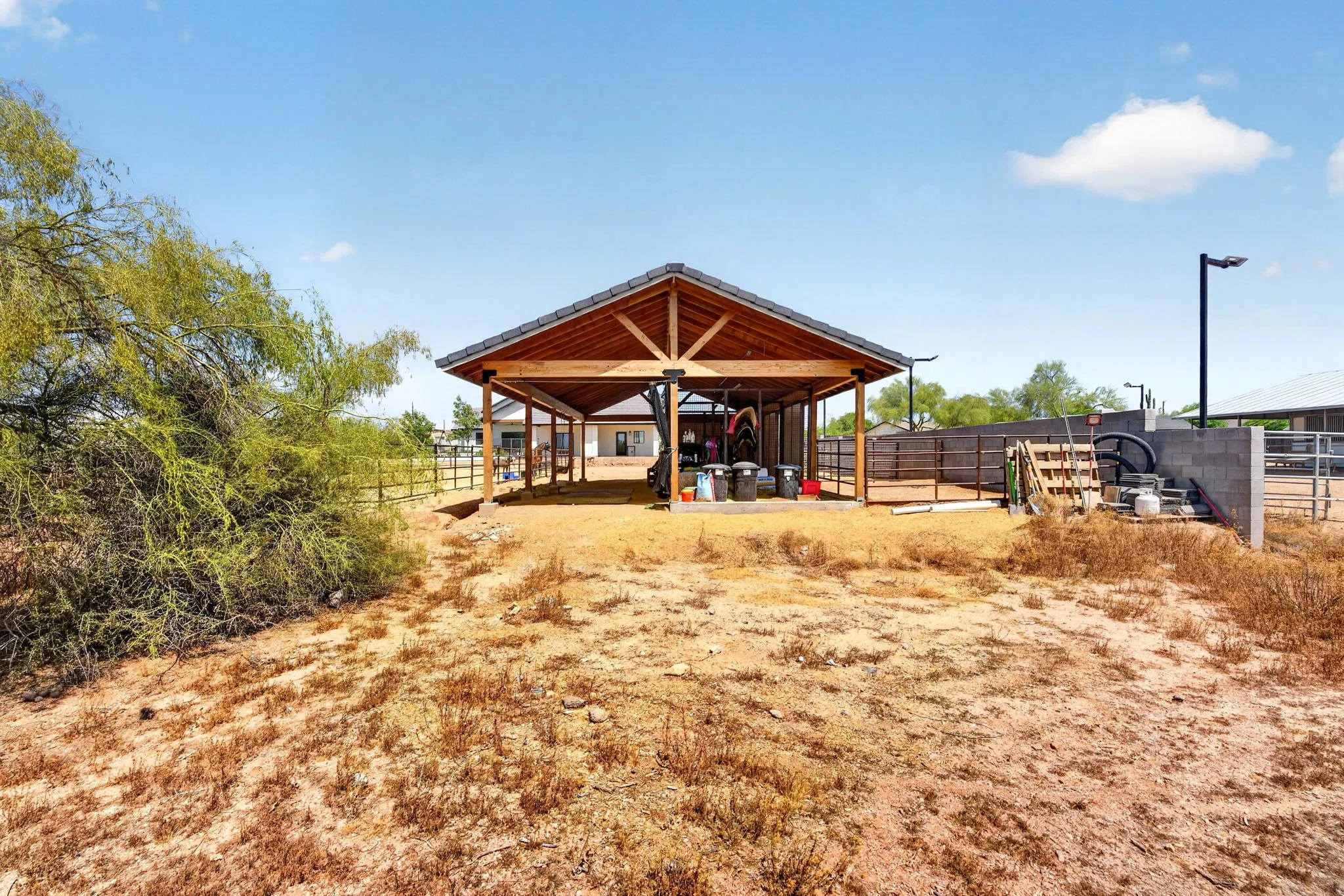 A wooden horse barn with a metal roof in a backyard under a blue sky with few clouds, surrounded by a fence, trees, and construction materials.