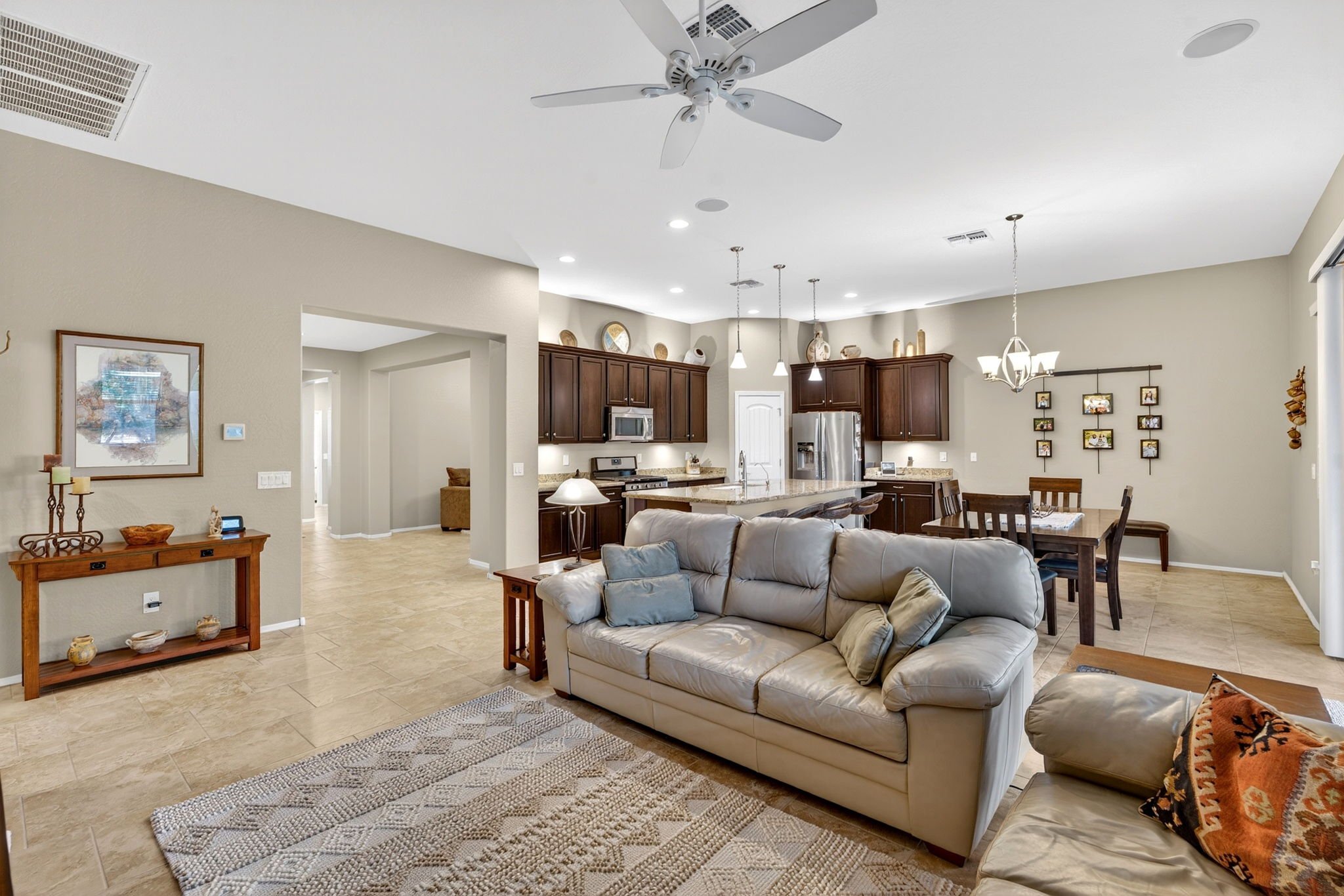 Open-concept living room and kitchen with beige tiled floor, cream walls, white ceiling, ceiling fan, and modern fixtures. The living area includes a beige sofa and decorative items on an adjacent wooden table. The kitchen features dark wooden cabine