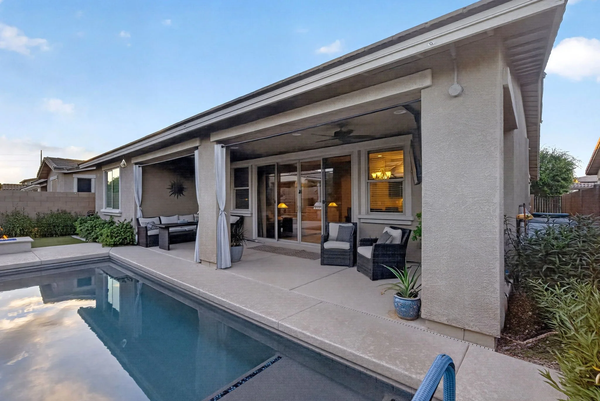 Backyard patio with a swimming pool, outdoor seating, potted plants, a covered area with curtains, and a house with large windows and a sliding door during sunset.