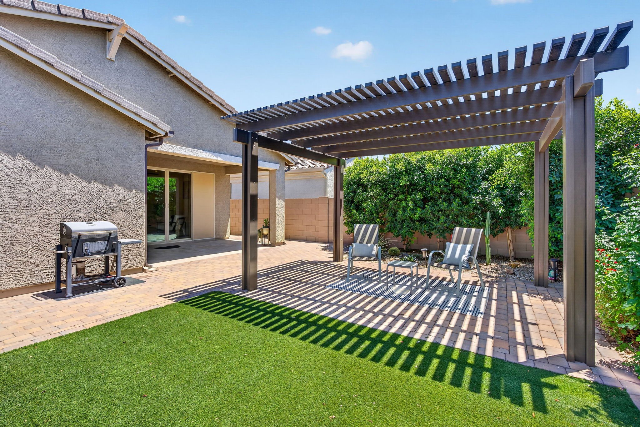 Backyard patio with a pergola, outdoor chairs, a green artificial lawn area, and lush trees and bushes.