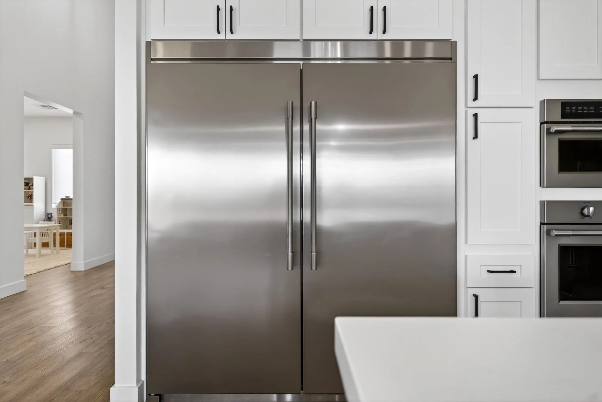 Stainless steel double refrigerator with black handles in a modern white kitchen with white cabinetry and black hardware.