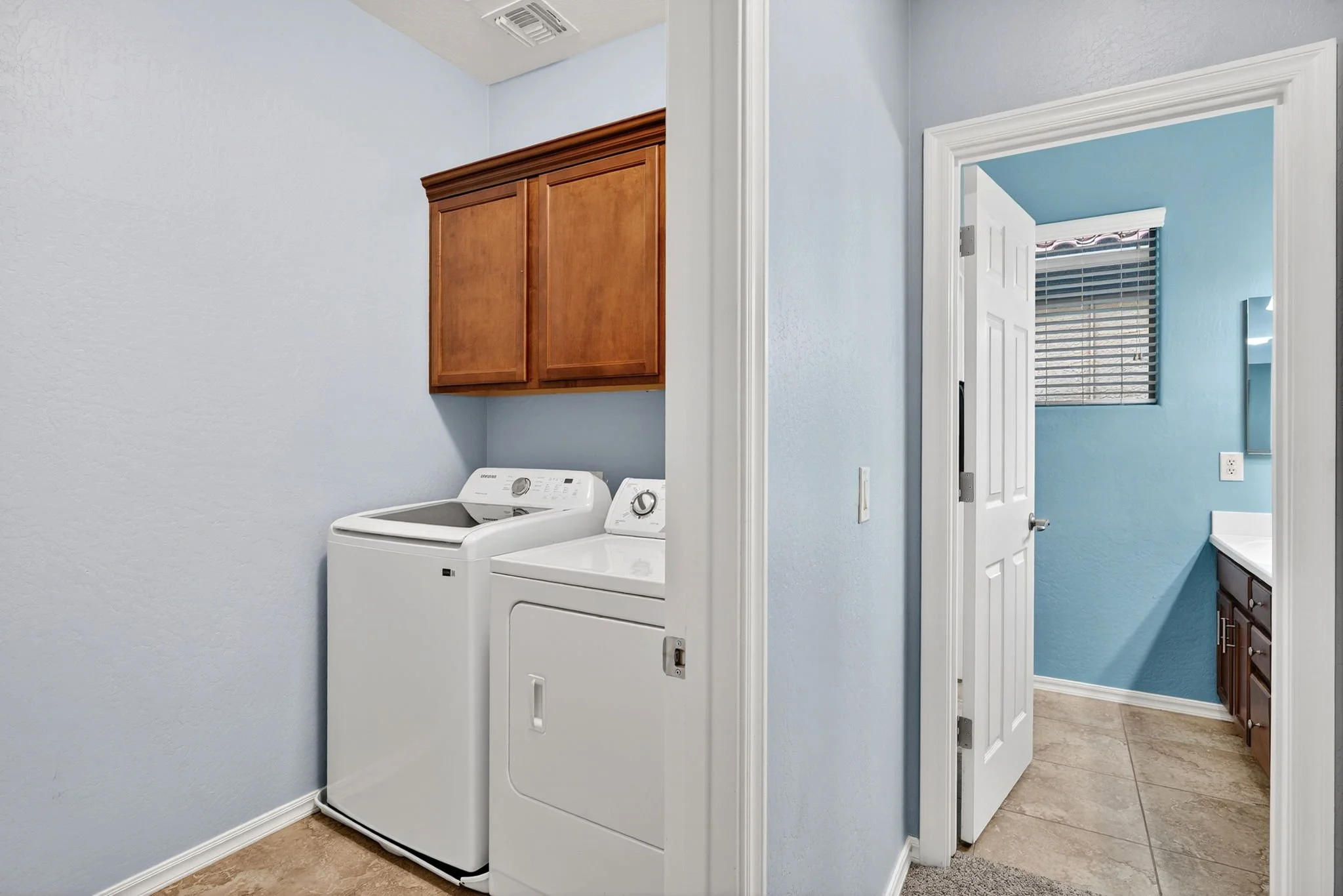 Laundry room with a white top-loading washing machine and a white dryer, light blue painted walls, a wooden cabinet above the machines, and a door opening to another room with a window and window blinds.