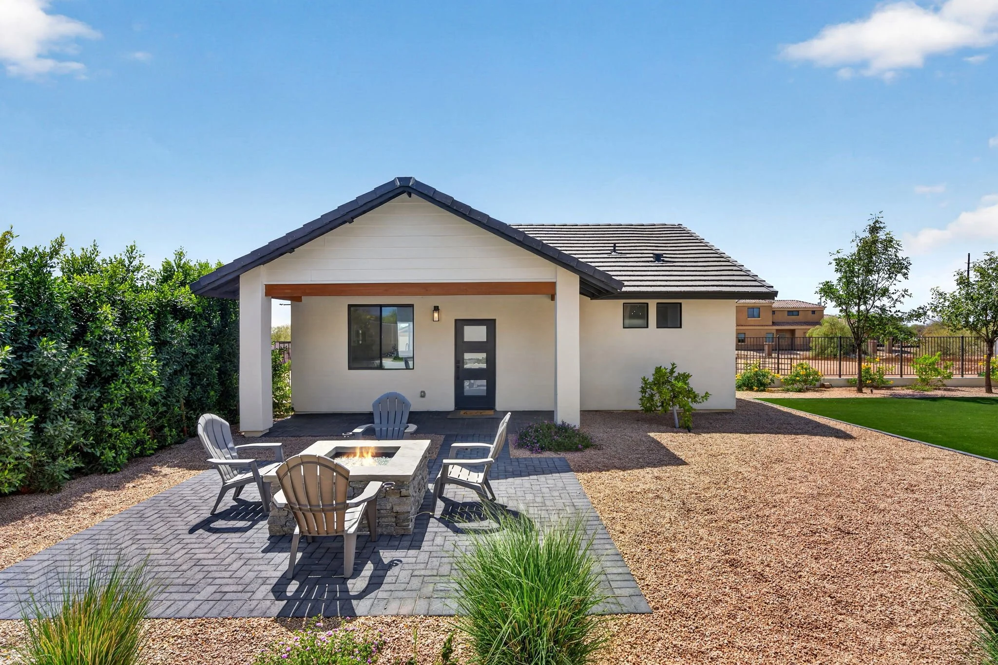 Backyard with a small white house, outdoor fire pit with chairs, paved patio area, bushes, trees, and a clear blue sky.