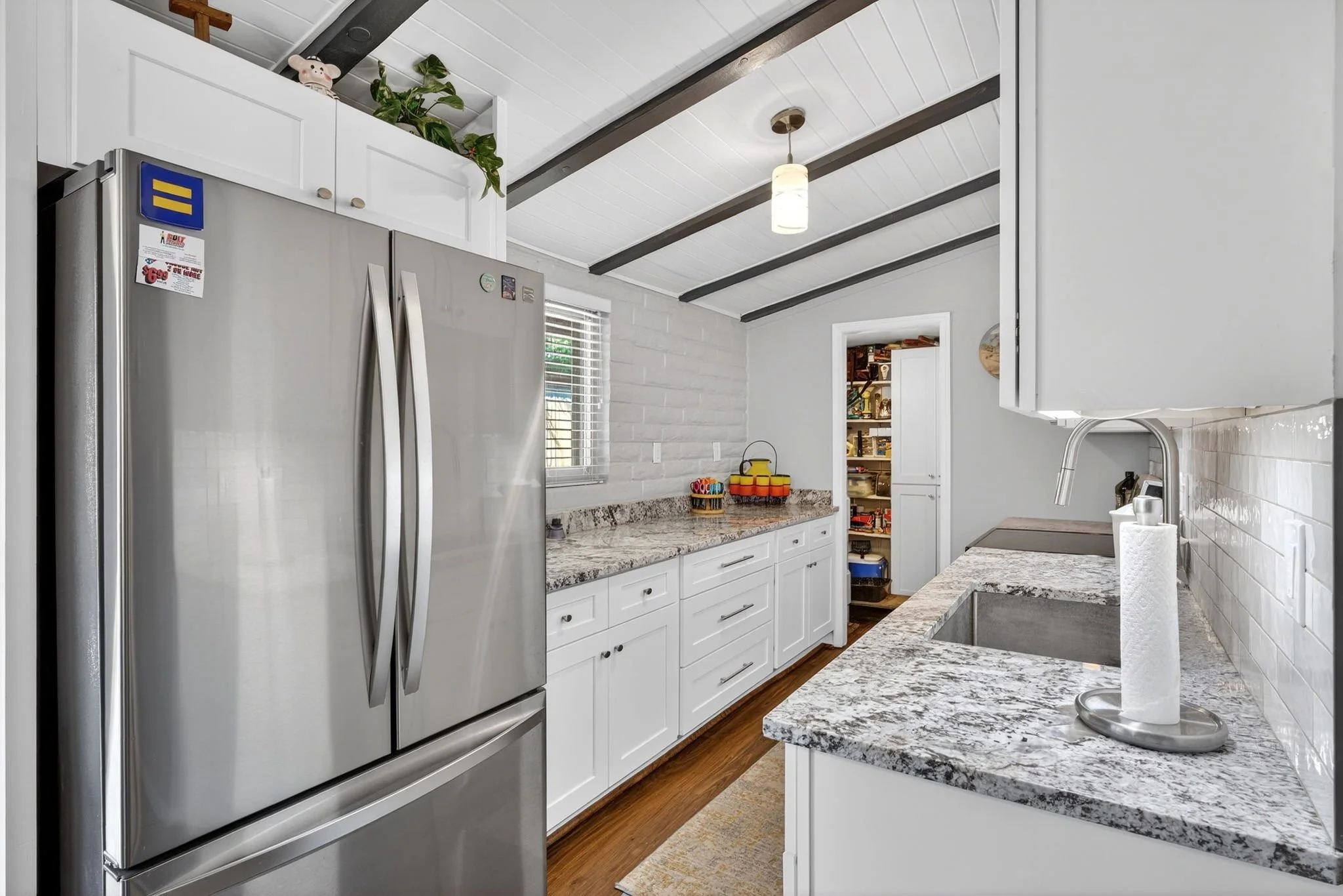 A modern kitchen with white cabinets, granite countertops, a stainless steel refrigerator, a small window with blinds, and a ceiling light. There is a paper towel roll on the counter and a pantry visible in the background.