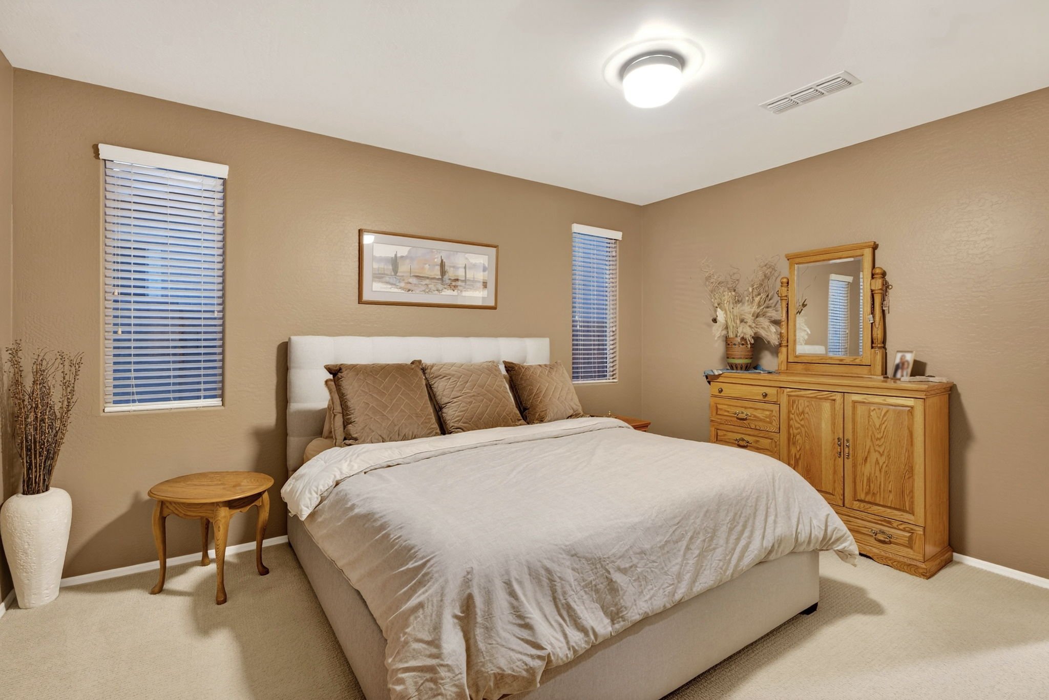 A bedroom with beige walls, a white bed with brown pillows, a round wooden side table, a large mirror on a wooden dresser, and two windows with blinds.