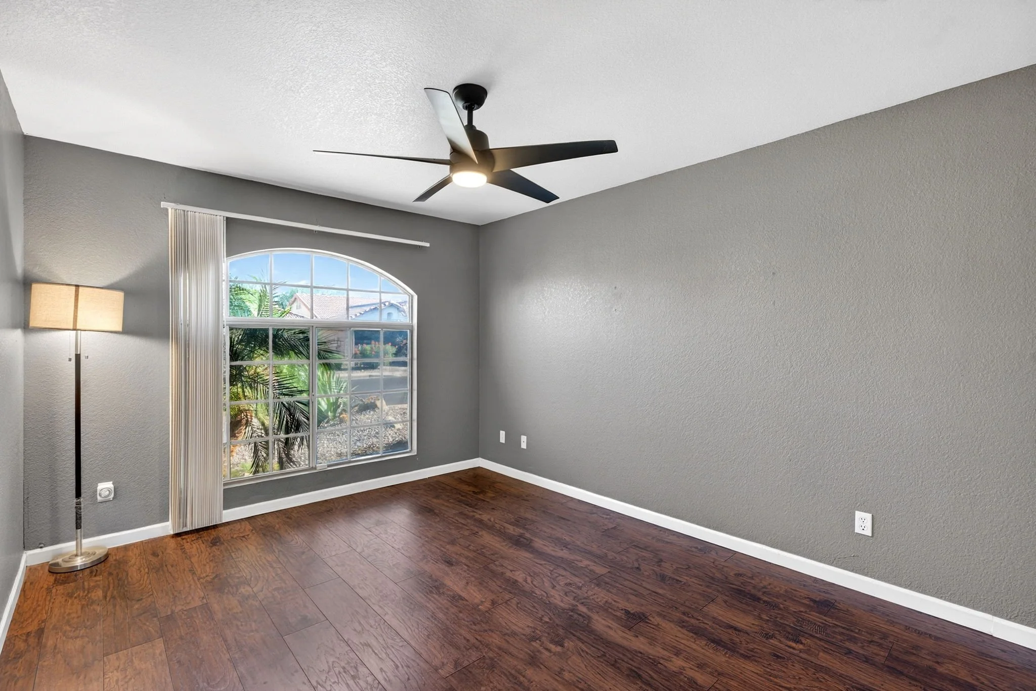Empty living room with gray walls, hardwood flooring, large window with vertical blinds, a ceiling fan with a light, and a standing lamp.