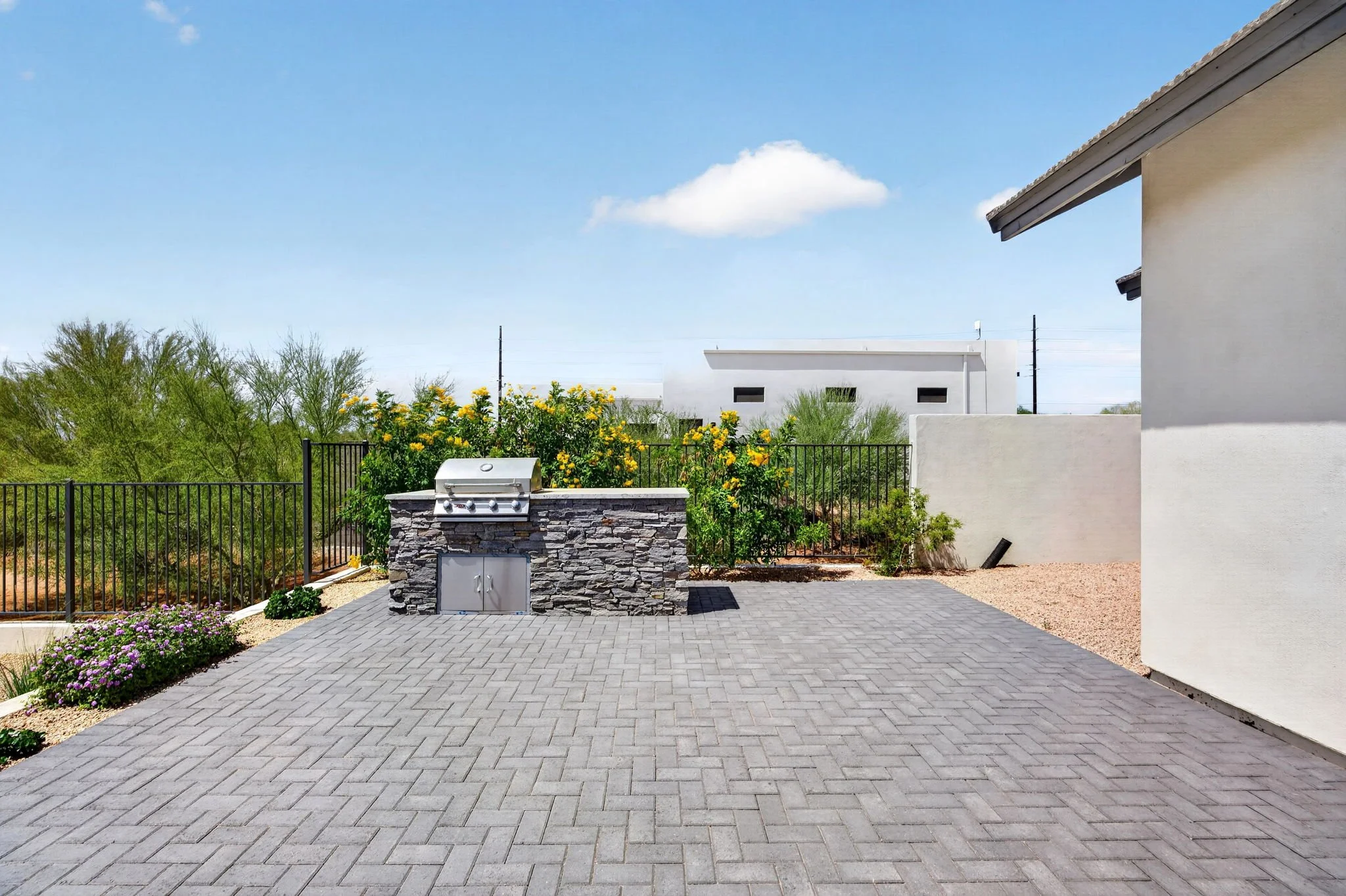 A backyard patio with gray pavers, a stone barbecue grill, and a black fence, with trees and white modern houses in the background. Sunny sky with a few clouds.