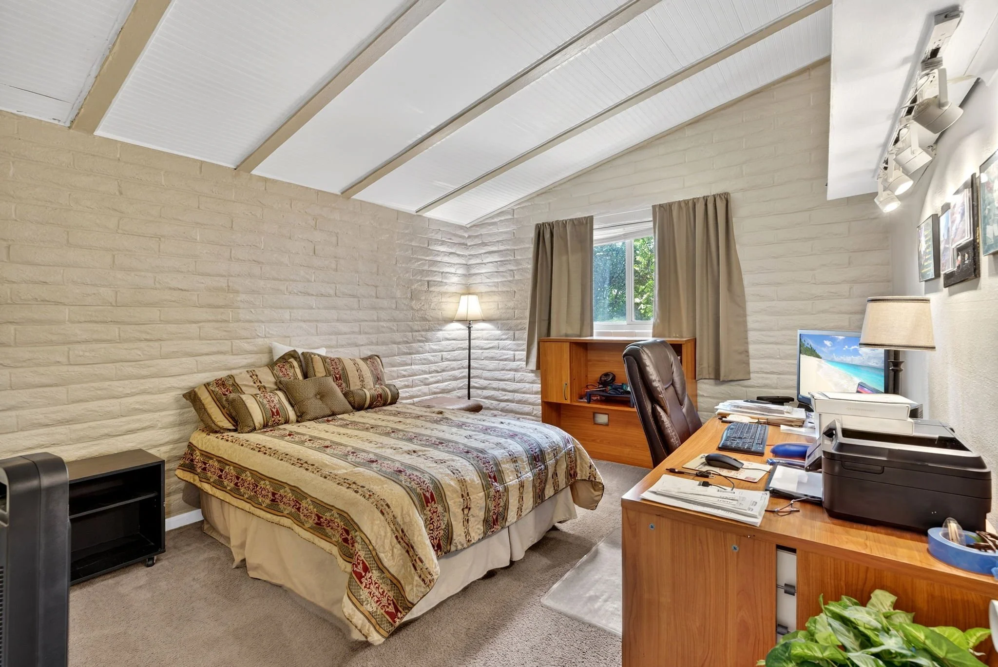 A bedroom with a bed, a window, and a desk with a computer, lamp, and office supplies.