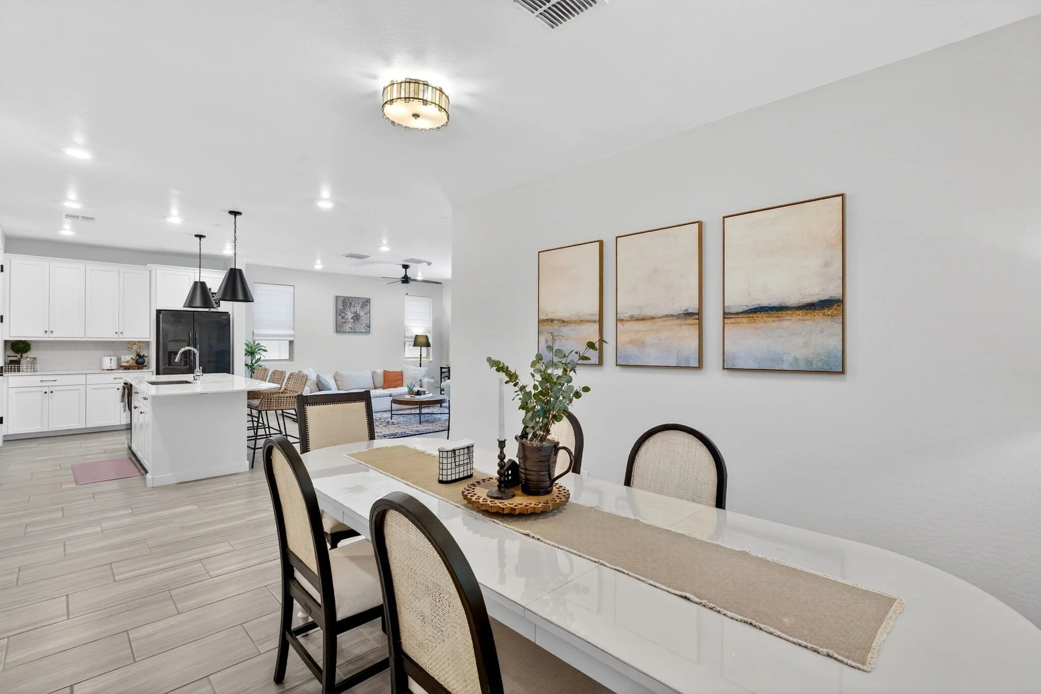 Open-concept living and dining area with white kitchen, black pendant lights, wooden dining table with black chairs, and wall art, decorated with a potted plant, with natural light coming through windows.