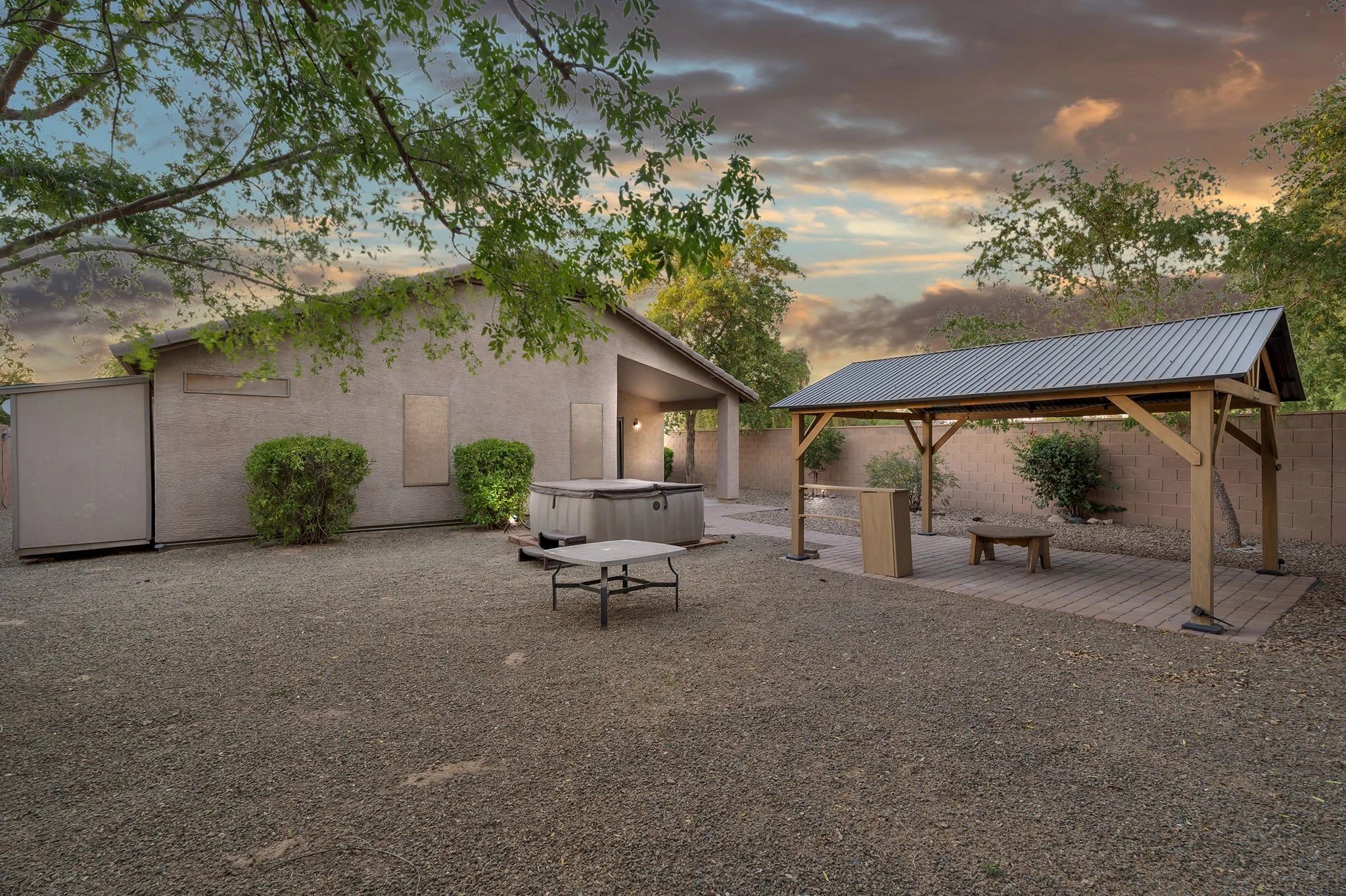 Backyard with gravel ground, two shrubs, a covered patio area with seating, a hot tub, and a building with a sloped roof, surrounded by a brick wall and trees at sunset.