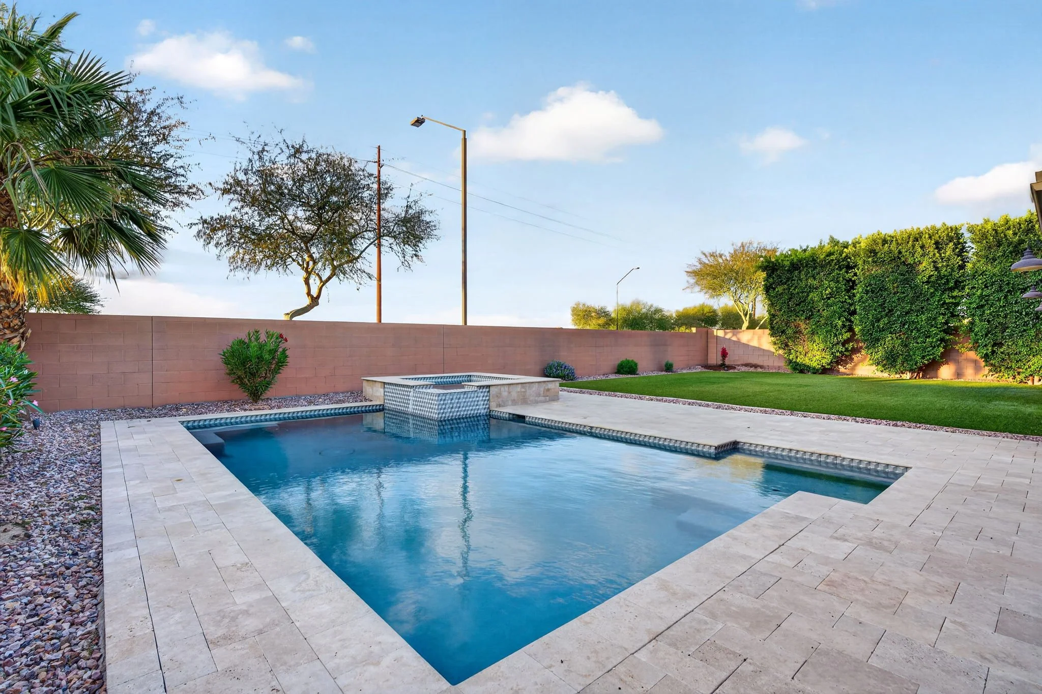 A backyard scene with a swimming pool surrounded by beige stone tiles, a brick wall, and green trees and bushes. There is a small fountain in the pool, and lamps on the right side. The sky is blue with some clouds.