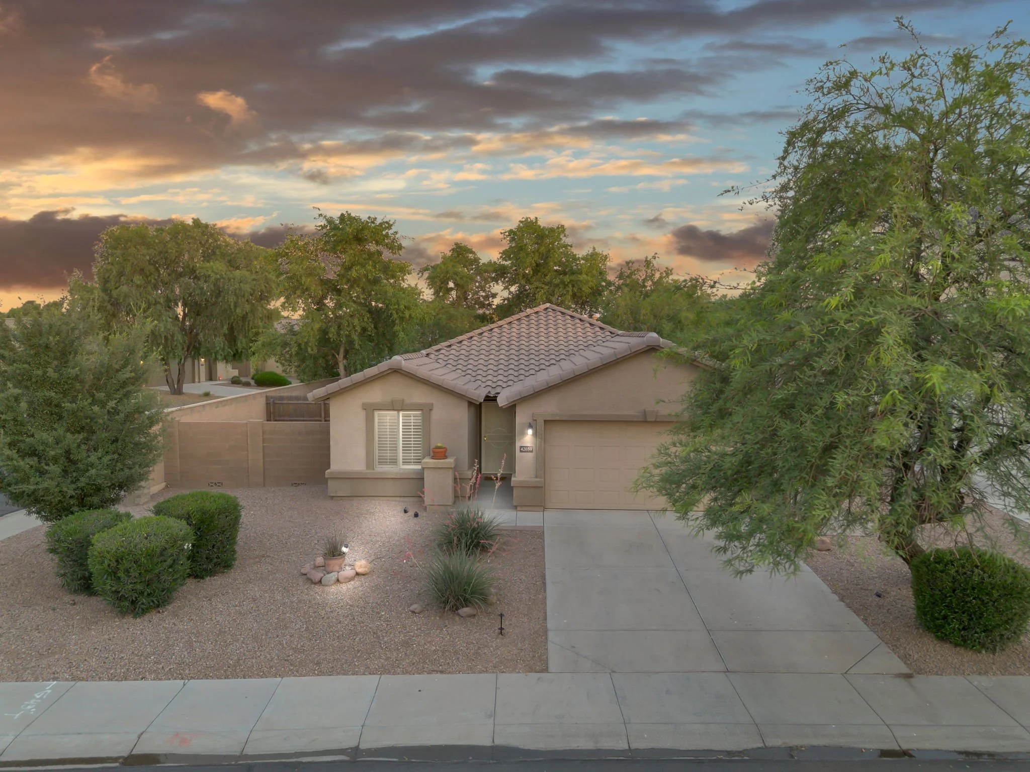 Front yard of a house with desert landscaping, a gravel ground cover, a small rock garden, and bushes. There are trees with green leaves and a house with a tiled roof and a garage. The sky is partly cloudy at dusk with orange and blue hues.
