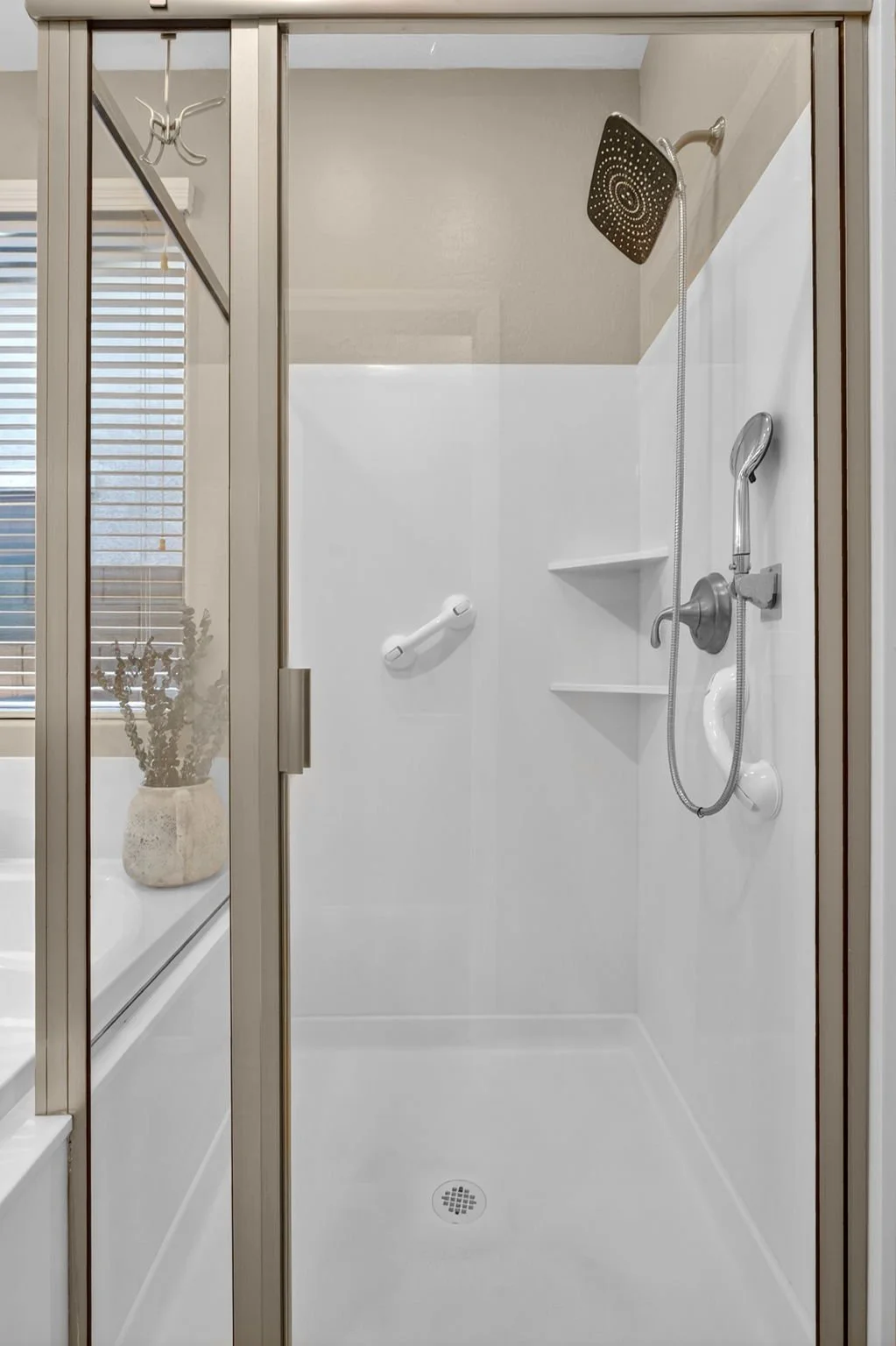 Empty walk-in shower with white walls, a showerhead, a handheld shower, a grab bar, and built-in shelves, enclosed by a glass door.