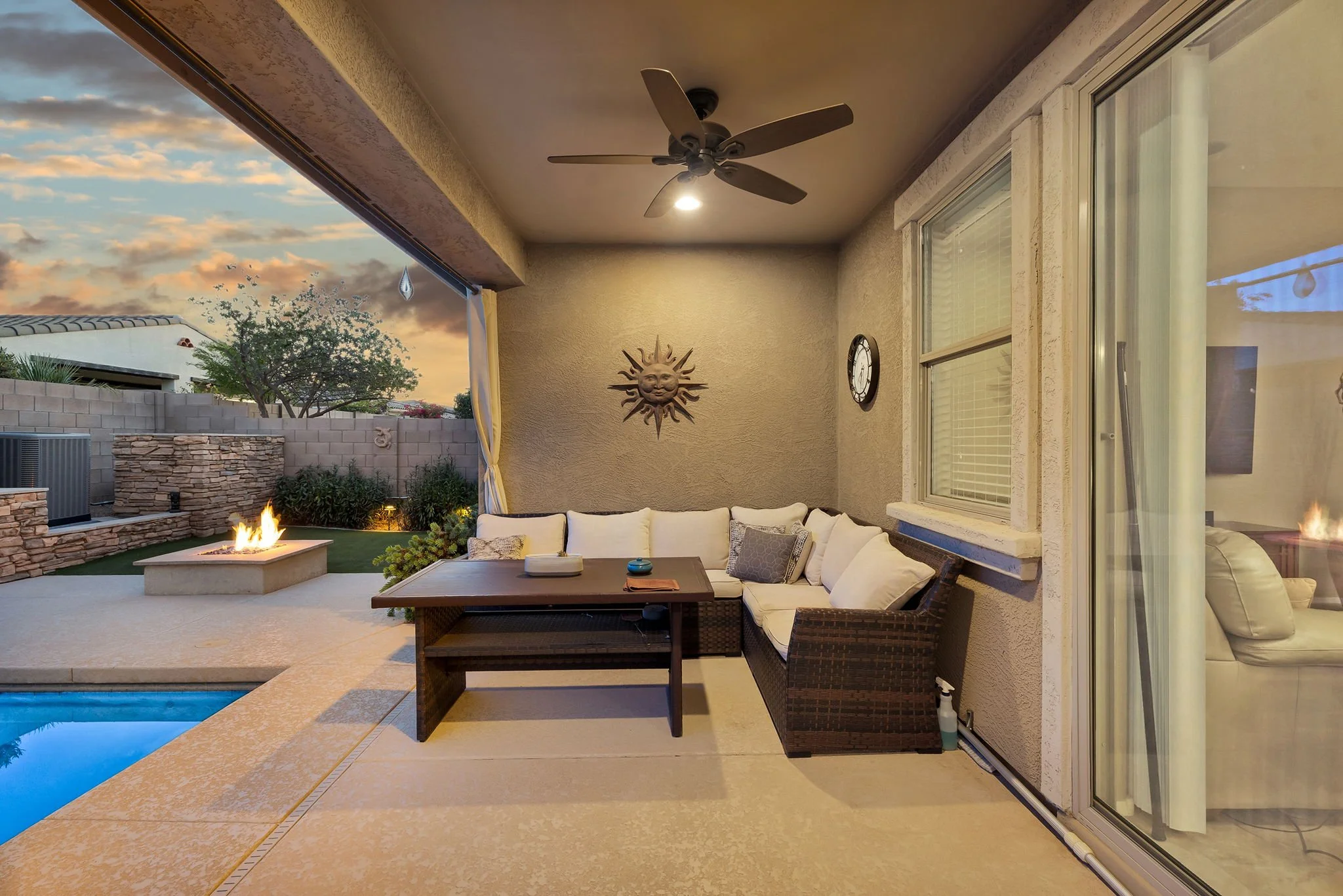 An outdoor patio area with a ceiling fan, wall clock, decorative sun face, and sliding glass doors leading inside. There is a comfortable beige sectional sofa with cushions, a coffee table, and a fire feature in the yard. The sky has a sunset with cl