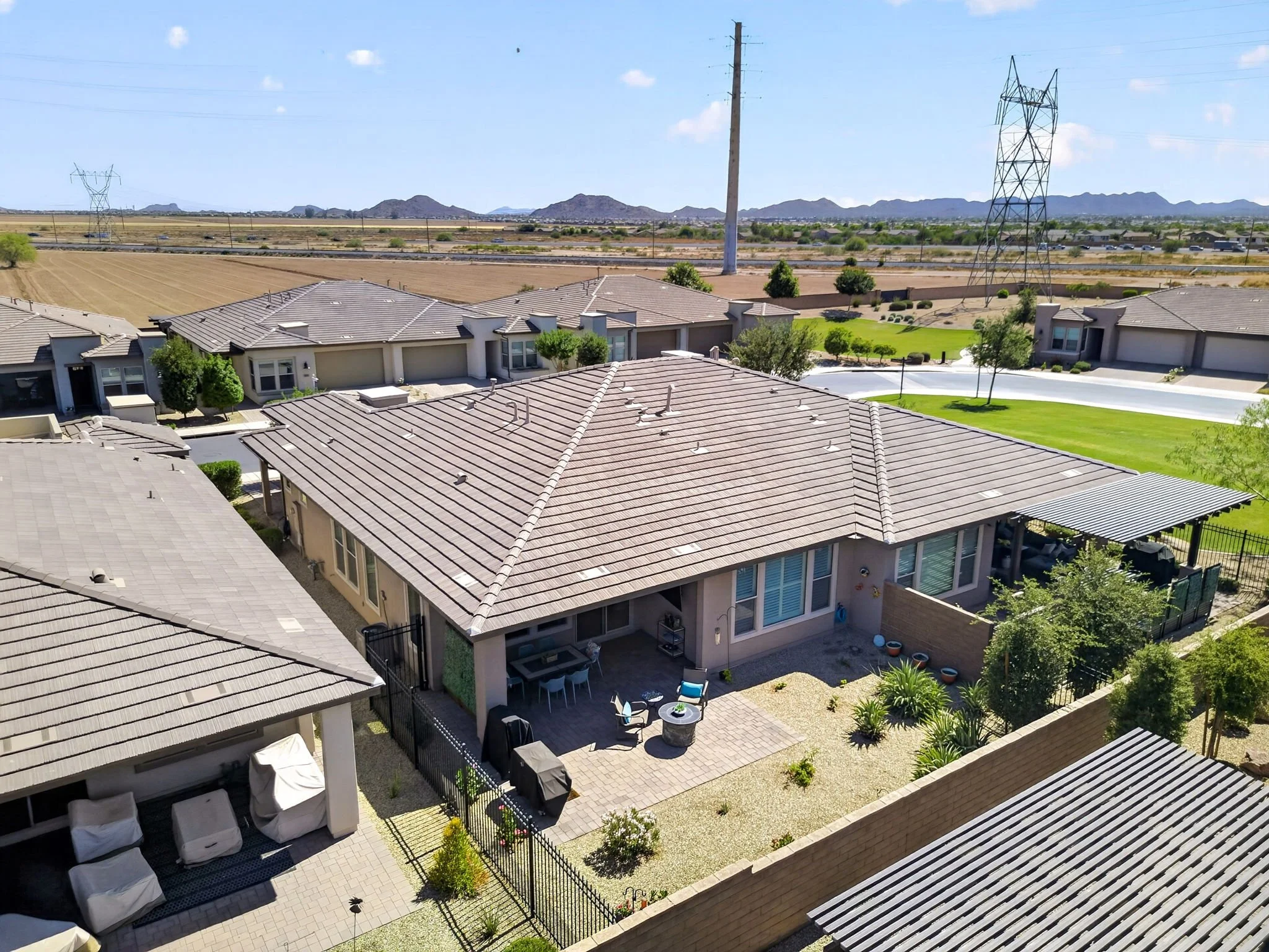 A view of a residential neighborhood with single-story houses, tiled roofs, and a backyard with patio furniture, green lawn, and trees. In the background, there are electrical power lines, transmission towers, desert landscape, and distant mountains 