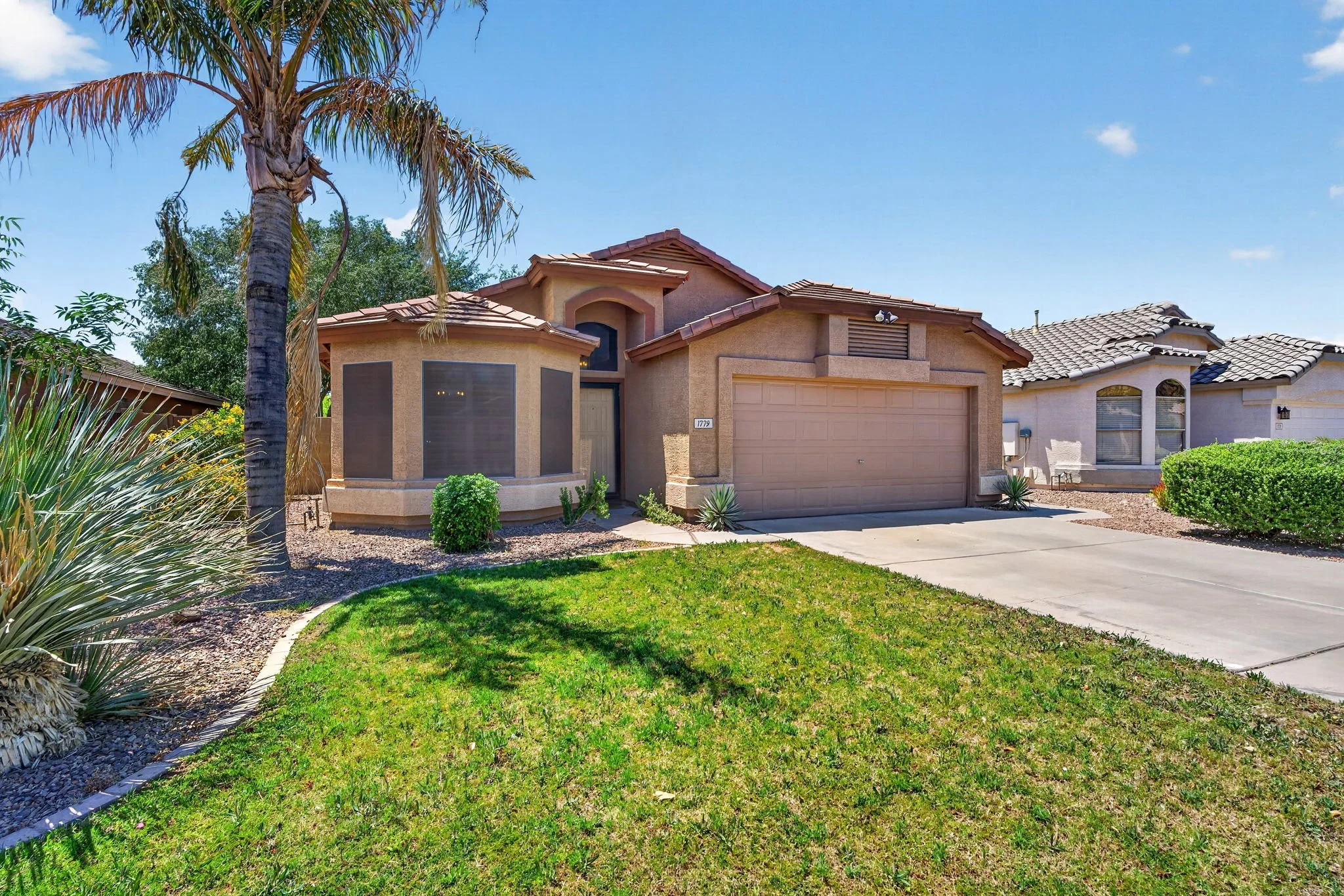 Front view of a two-story house with a brown garage door, beige exterior, and a landscaped yard with green grass, bushes, and a palm tree, under a clear blue sky.