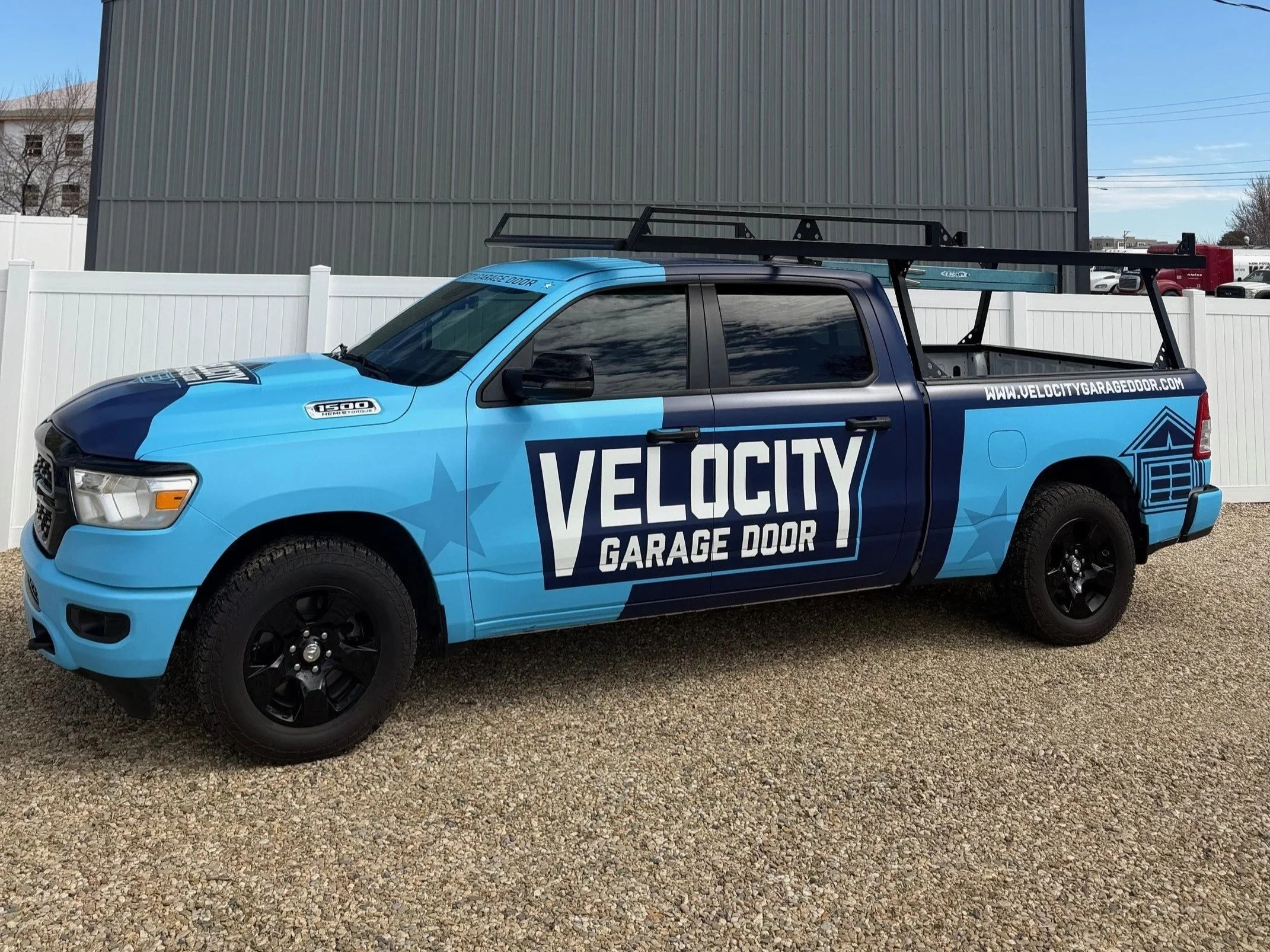 A blue pickup truck with branding for Velocity Garage Door parked on gravel, with a white fence and a large gray building in the background.