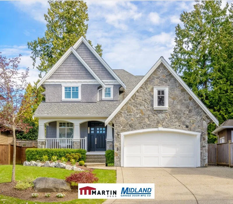 A two-story house with a stone and grey siding exterior, white garage door, and front porch with white railing, surrounded by landscaped yard and tall green trees.