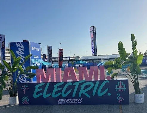 Sign reading 'Miami Electric' placed in front of a pool area with plants and tall banners on a sunny day.