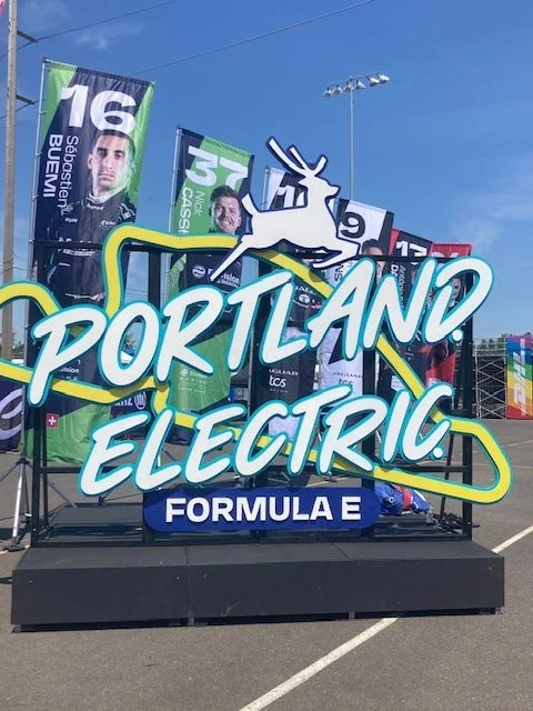 Stage with a sign reading 'Portland Electric Formula E' and flags with racing driver images, set outdoors at a racing event.