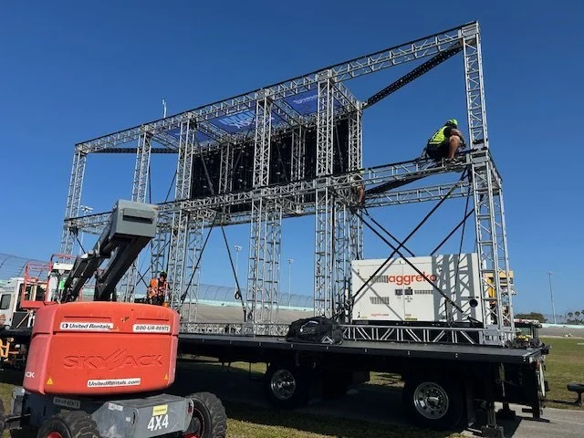 Construction workers assembling a large steel frame structure on a flatbed truck, with a blue sky and a crane nearby.