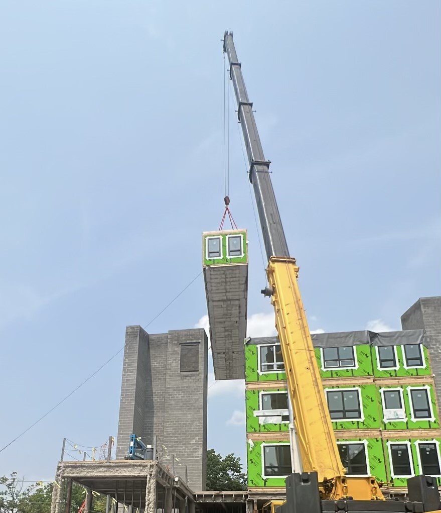 A construction crane lifting a modular building unit onto a partially constructed green building.