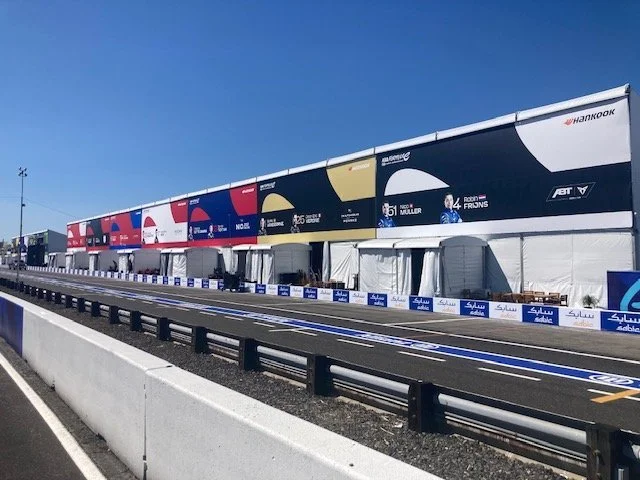 Long building with colorful banners and white tents along a race track under a clear blue sky.