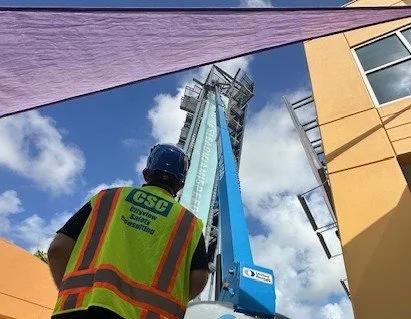 A construction worker wearing a safety vest and helmet looking up at a tall construction crane during the day.
